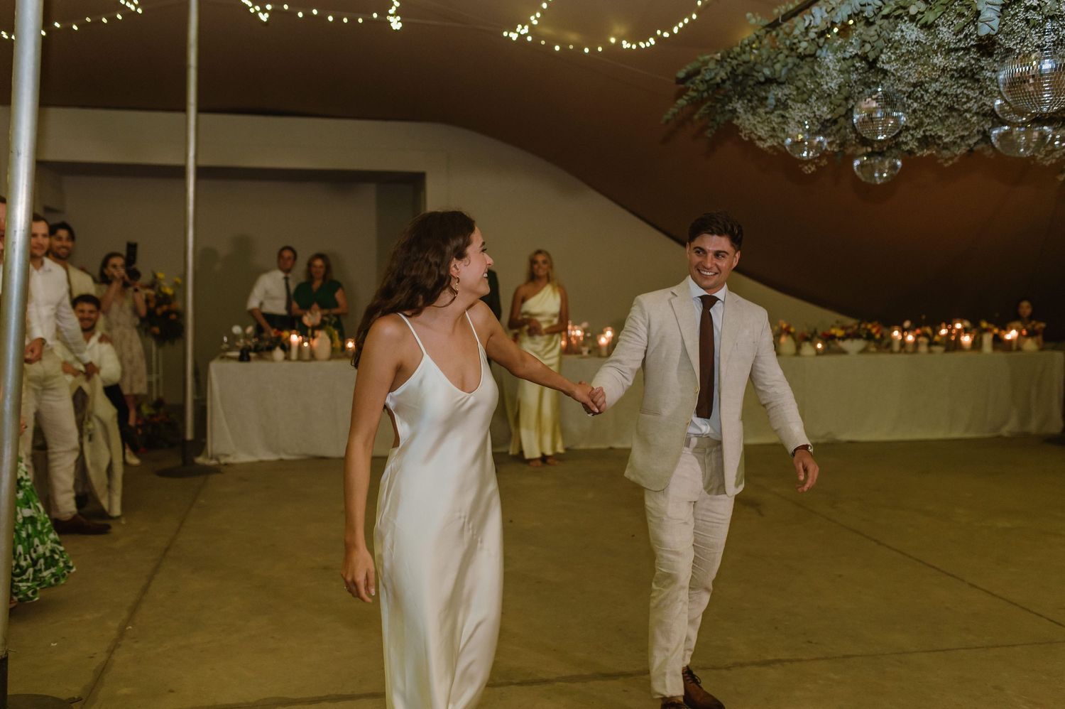 A couple enters their wedding reception under twinkling lights in a tent venue.