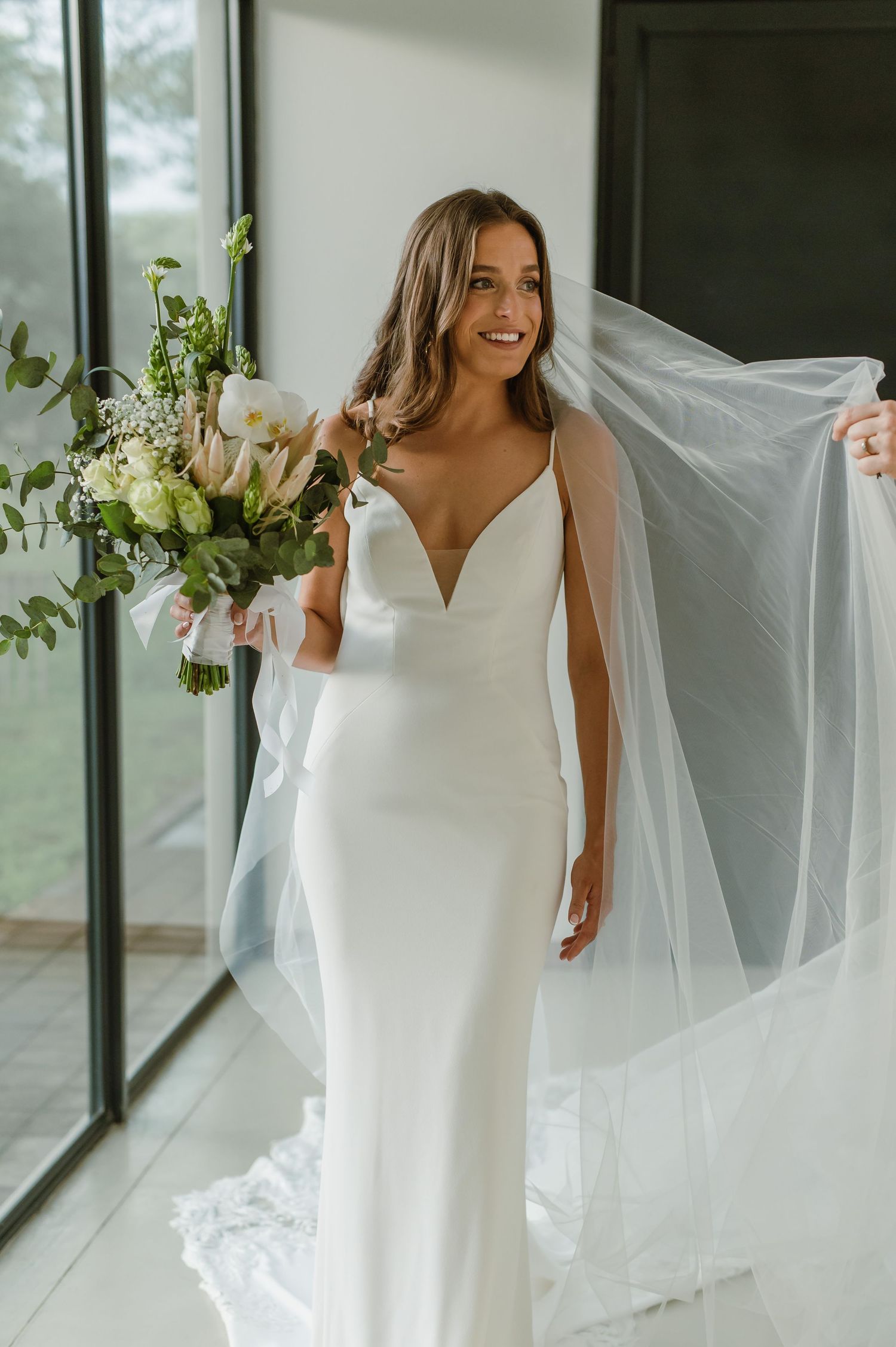 Bride in sleek white gown twirling with flowing veil in modern indoor setting.