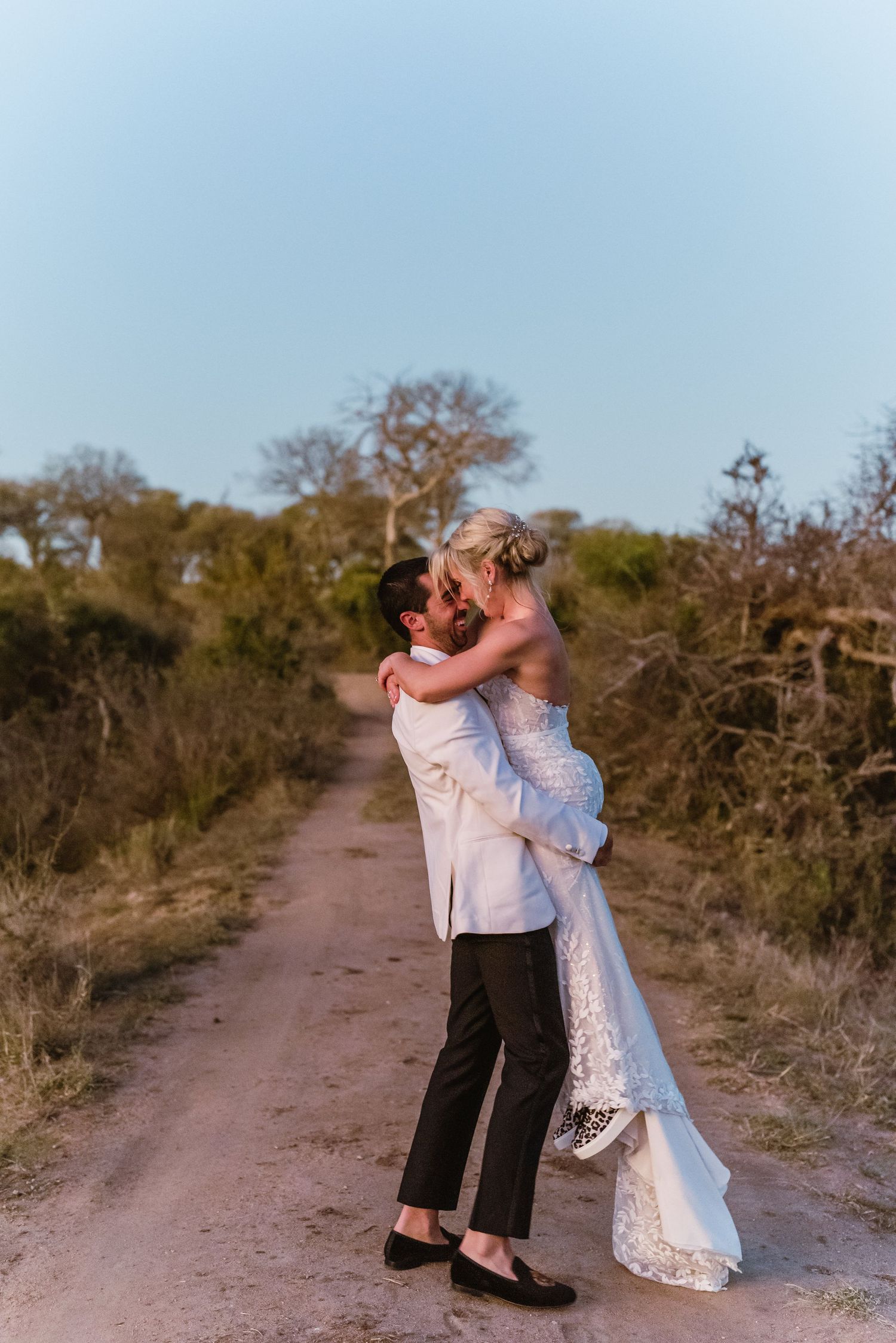 Romantic couple embraces on a rustic dirt path surrounded by trees at sunset during their wedding photo session.