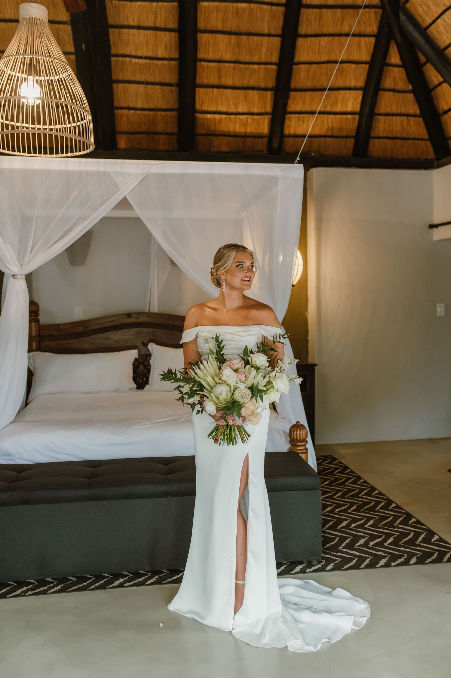 Bride in sleek white gown with thigh slit holds luxurious bouquet under thatched roof.