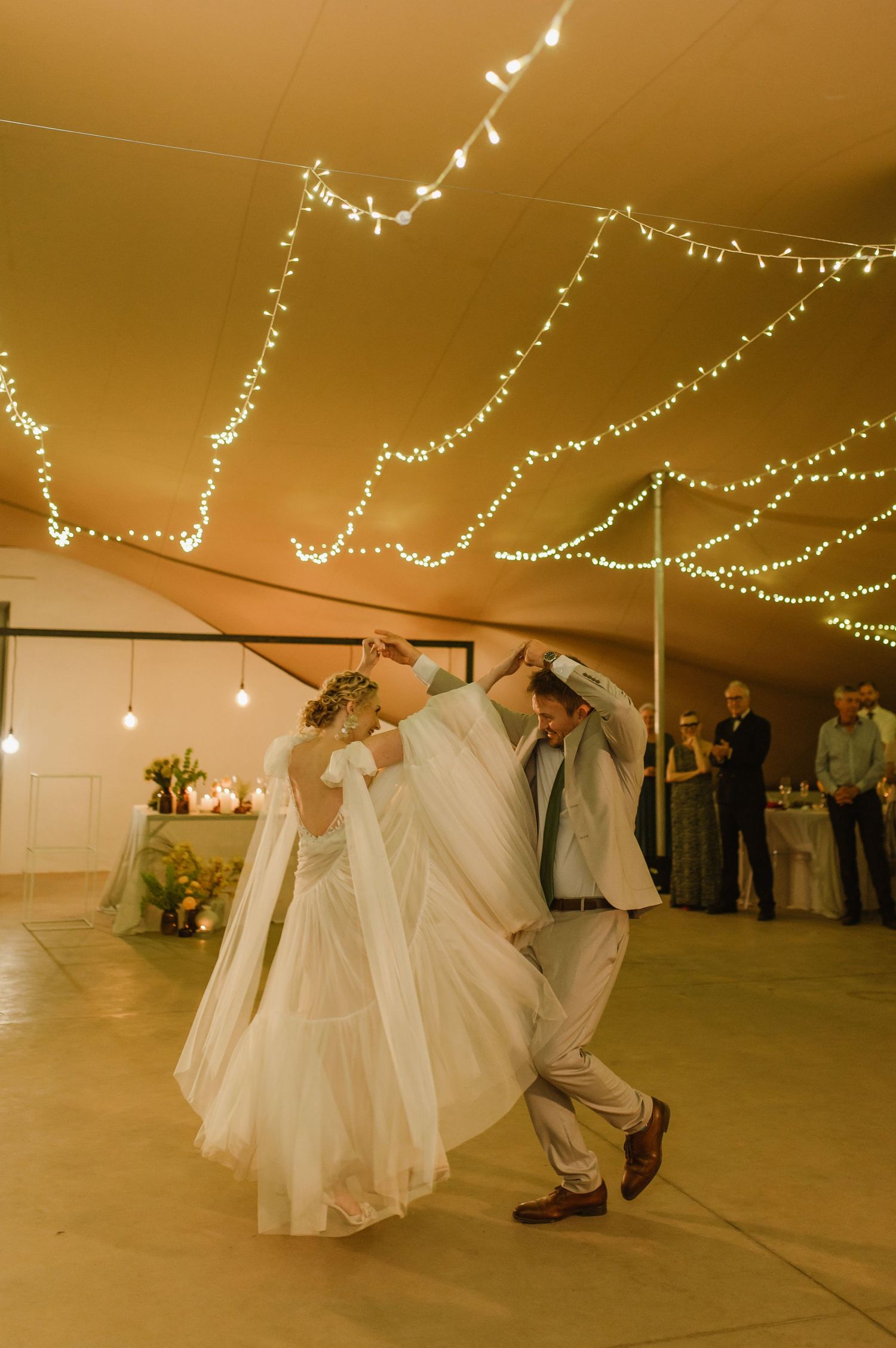 Wedding couple dances under warm string lights in an elegant reception tent.
