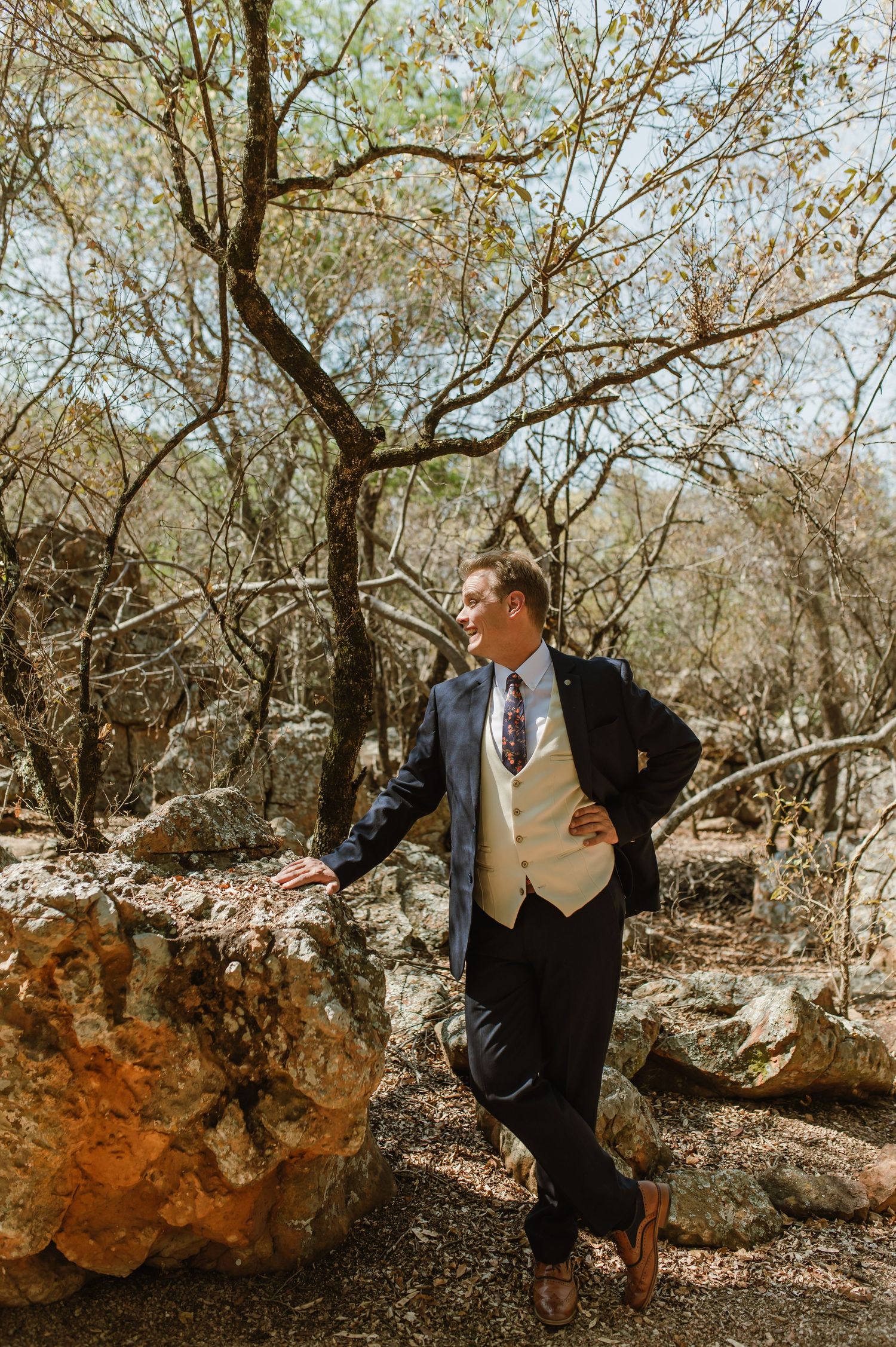 A suited figure leans against a rock in a wooded outdoor setting wearing formal attire.