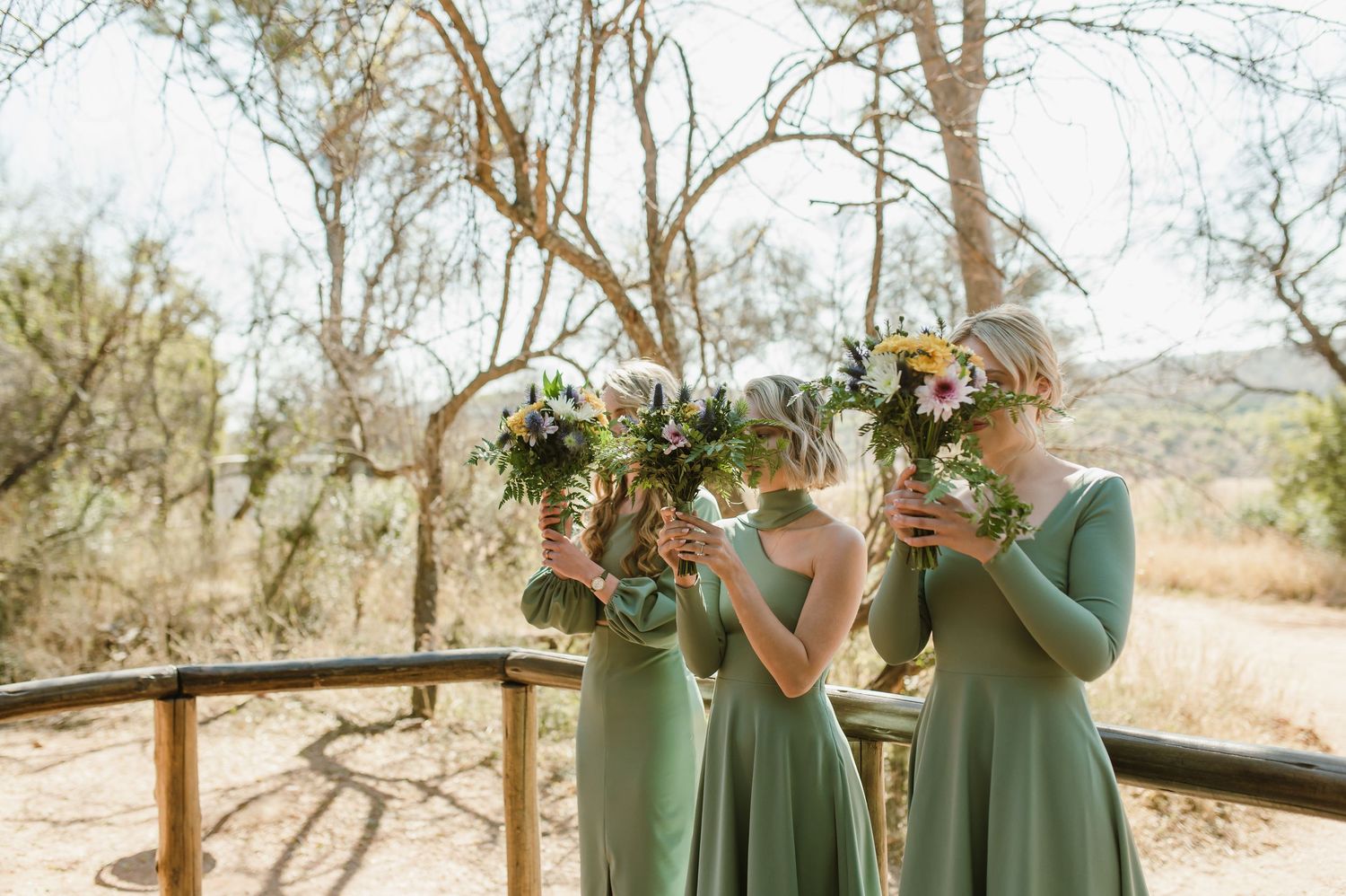 Bridesmaids in sage green dresses holding yellow and white bouquets outdoors.
