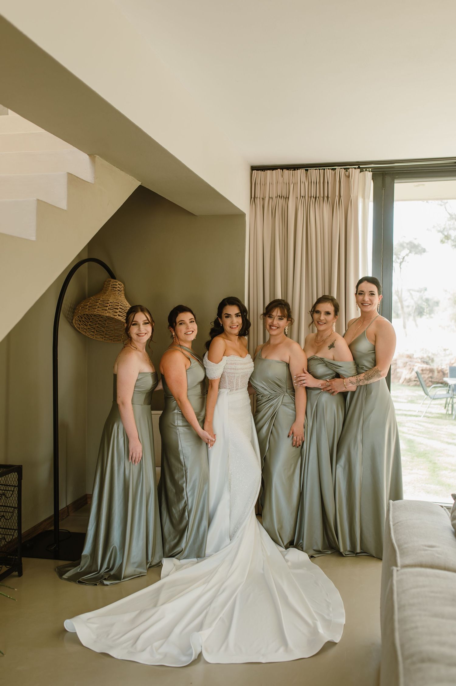 Wedding party in sage green dresses poses elegantly against a white wall.