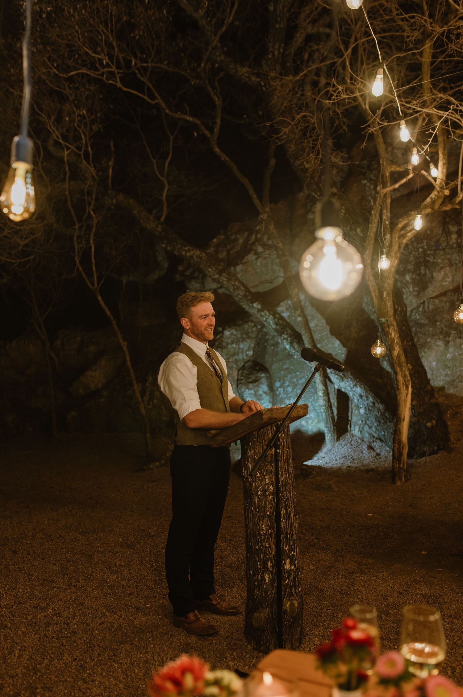 String lights illuminate a rustic outdoor dining setup with red floral arrangements.