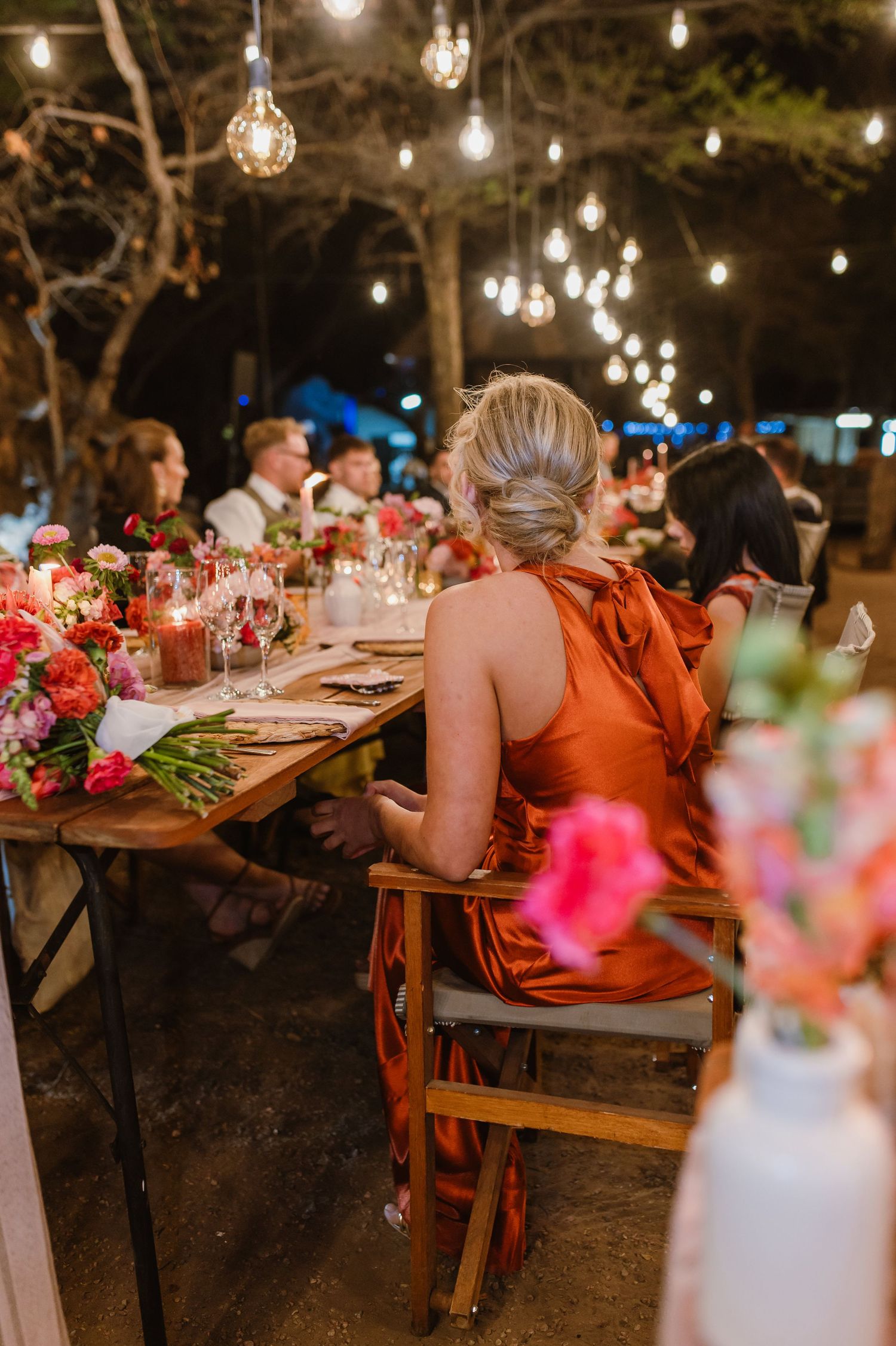 Evening outdoor dining setup with hanging lights and floral centerpieces on long wooden tables.