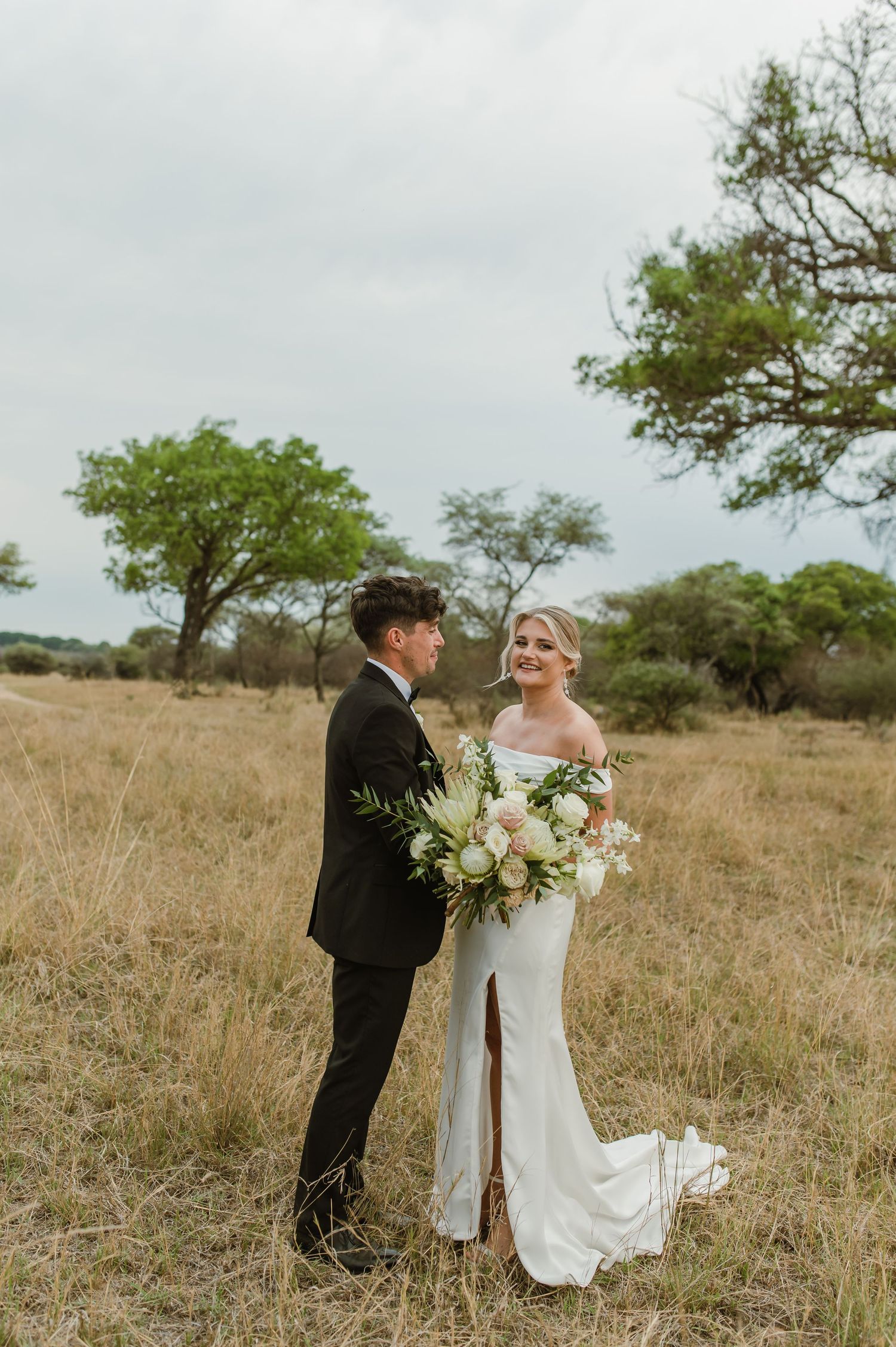 A wedding couple poses together in an open field with scattered trees on an overcast day.