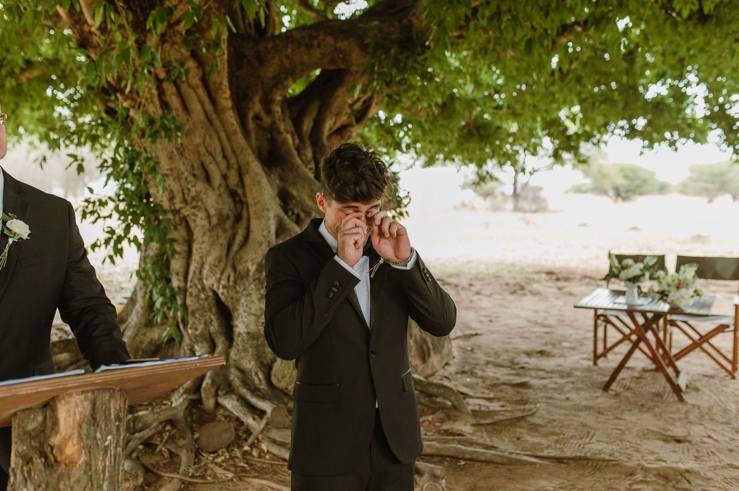 Guests in formal attire gather under a large tree during outdoor wedding ceremony.