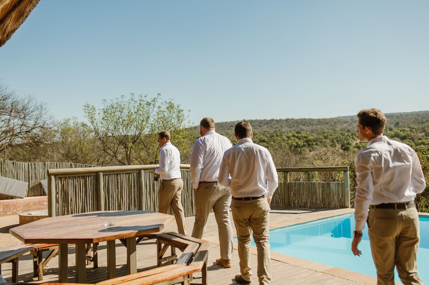 Groomsmen gather on a deck overlooking a swimming pool at a scenic outdoor venue.