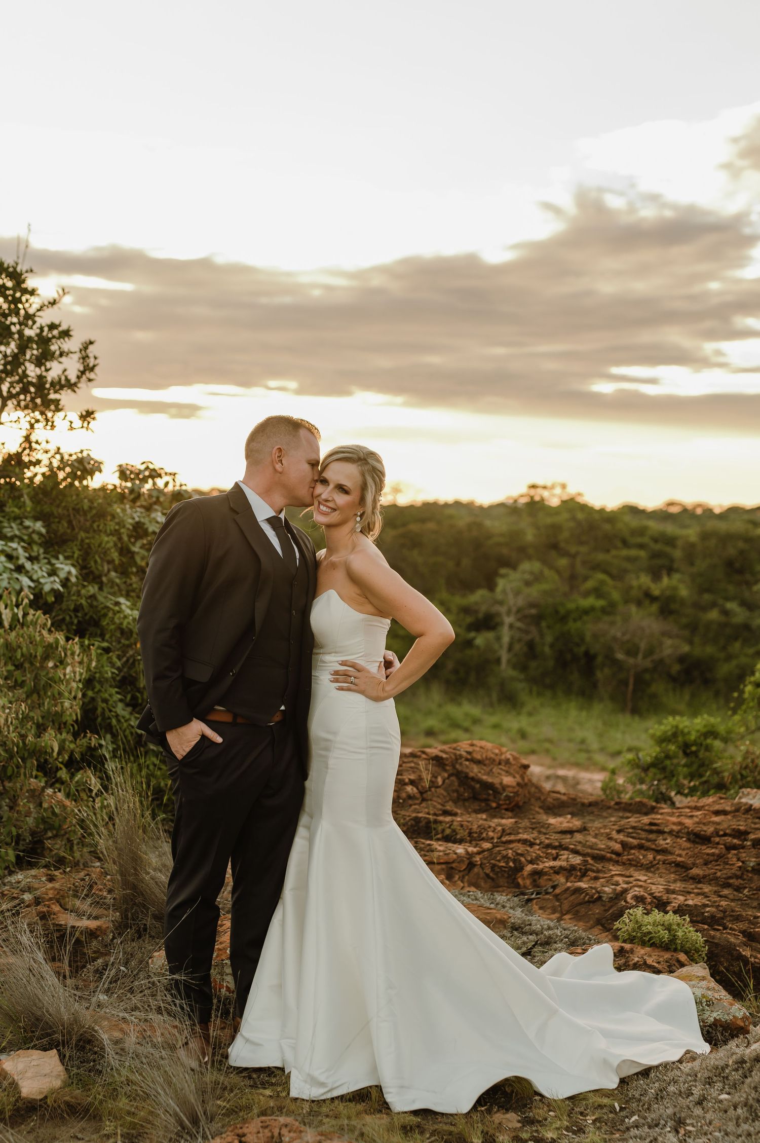 Bride in flowing white gown and groom pose on rocky terrain during sunset for wedding photos.