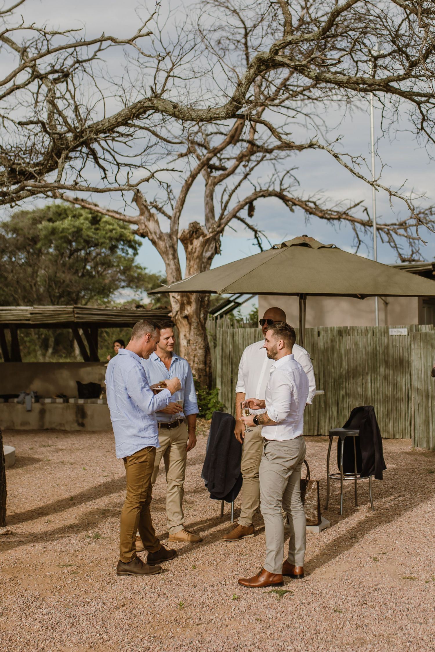 Group of people mingling at an outdoor gathering under trees on gravel ground.