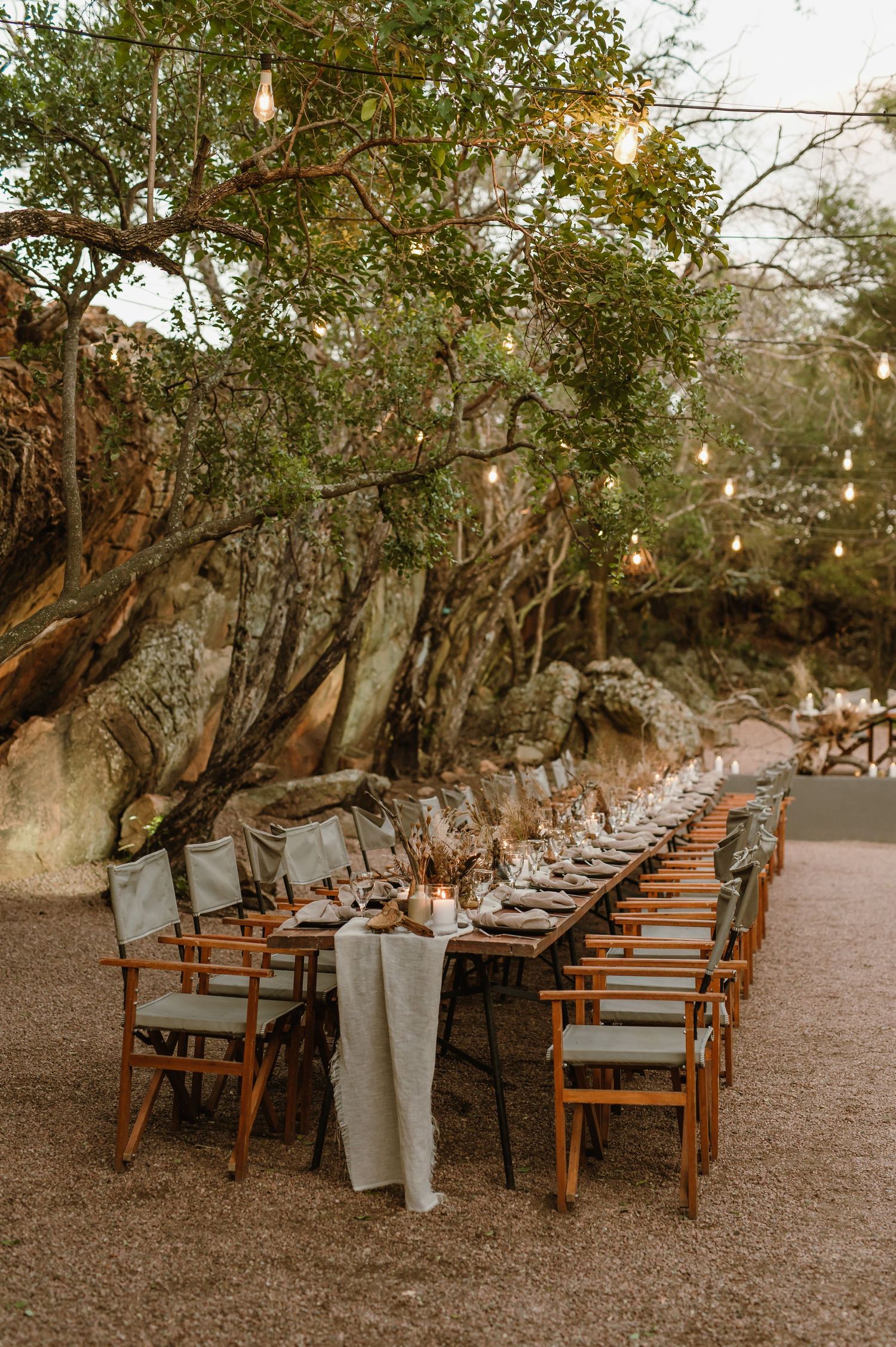 Outdoor wedding reception setup with wooden chairs and tables arranged under natural tree canopy.