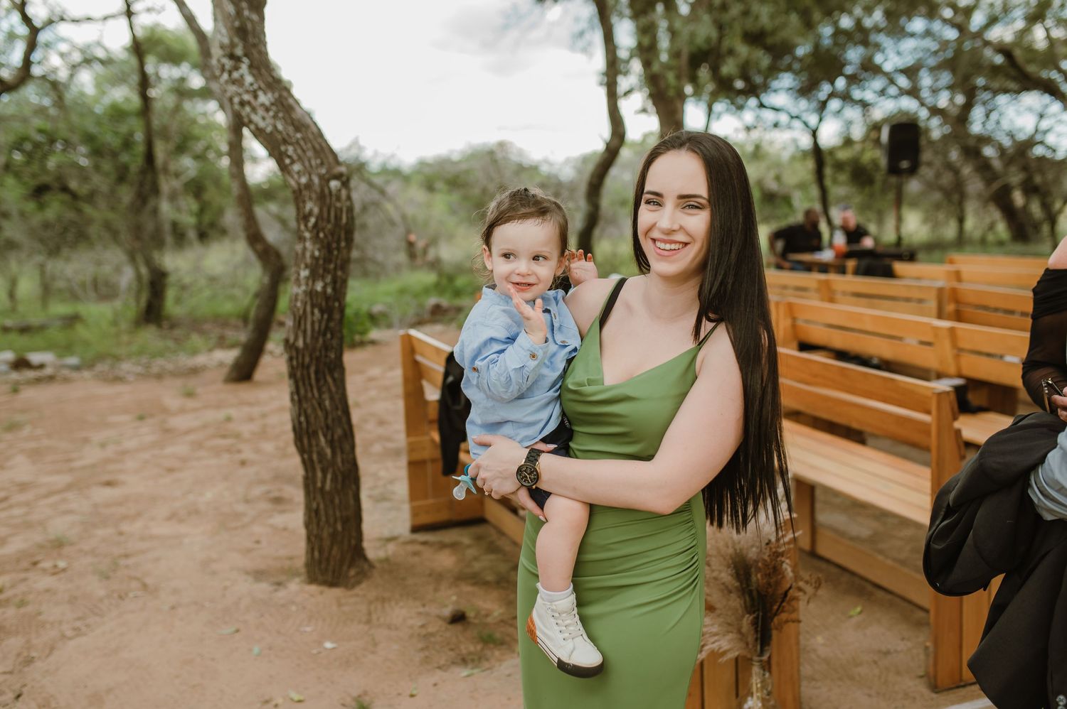 A guest in a green dress holds a small child at an outdoor wedding venue with wooden benches.