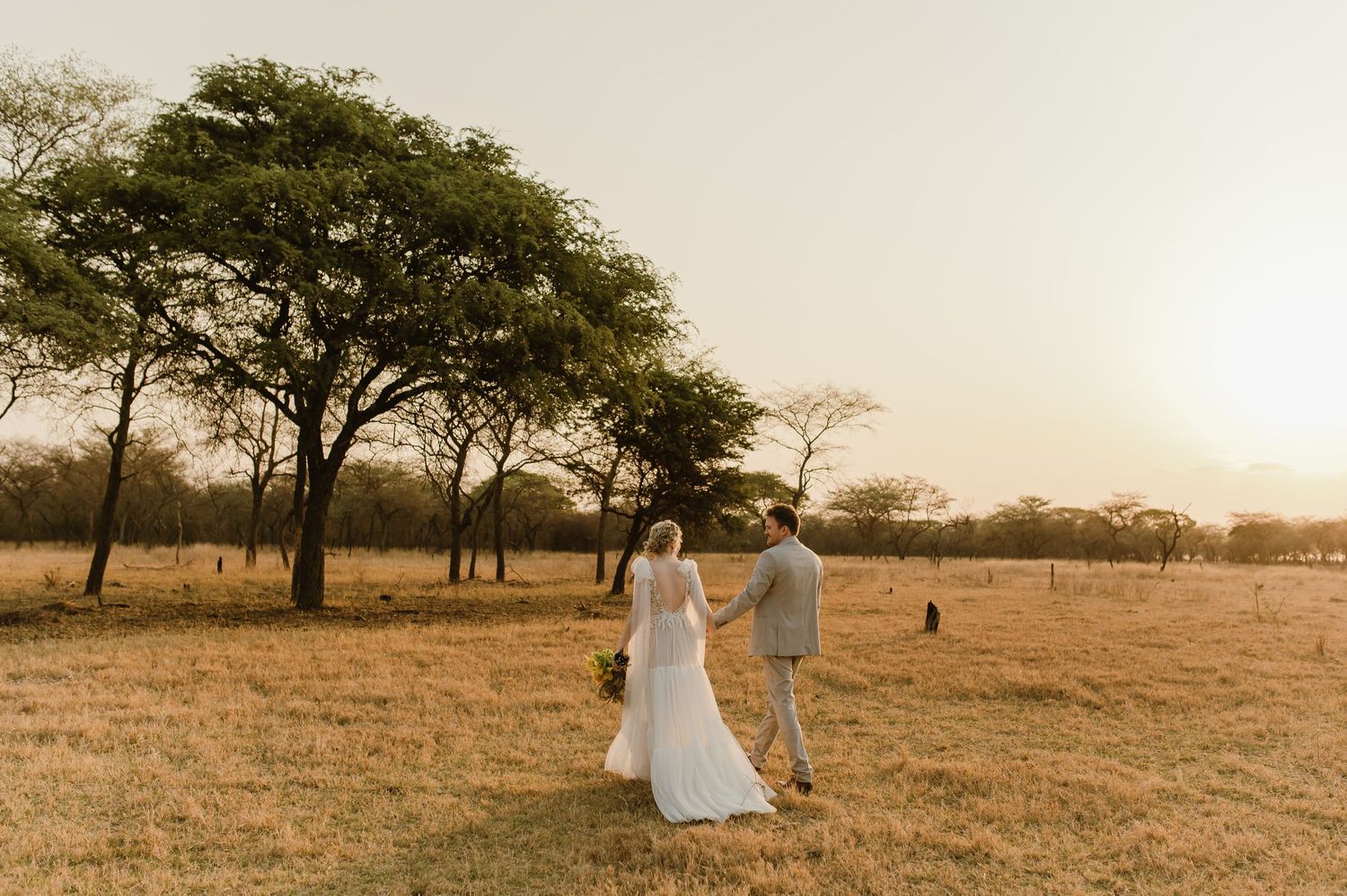Couple walking through savanna grassland with acacia trees at sunset.
