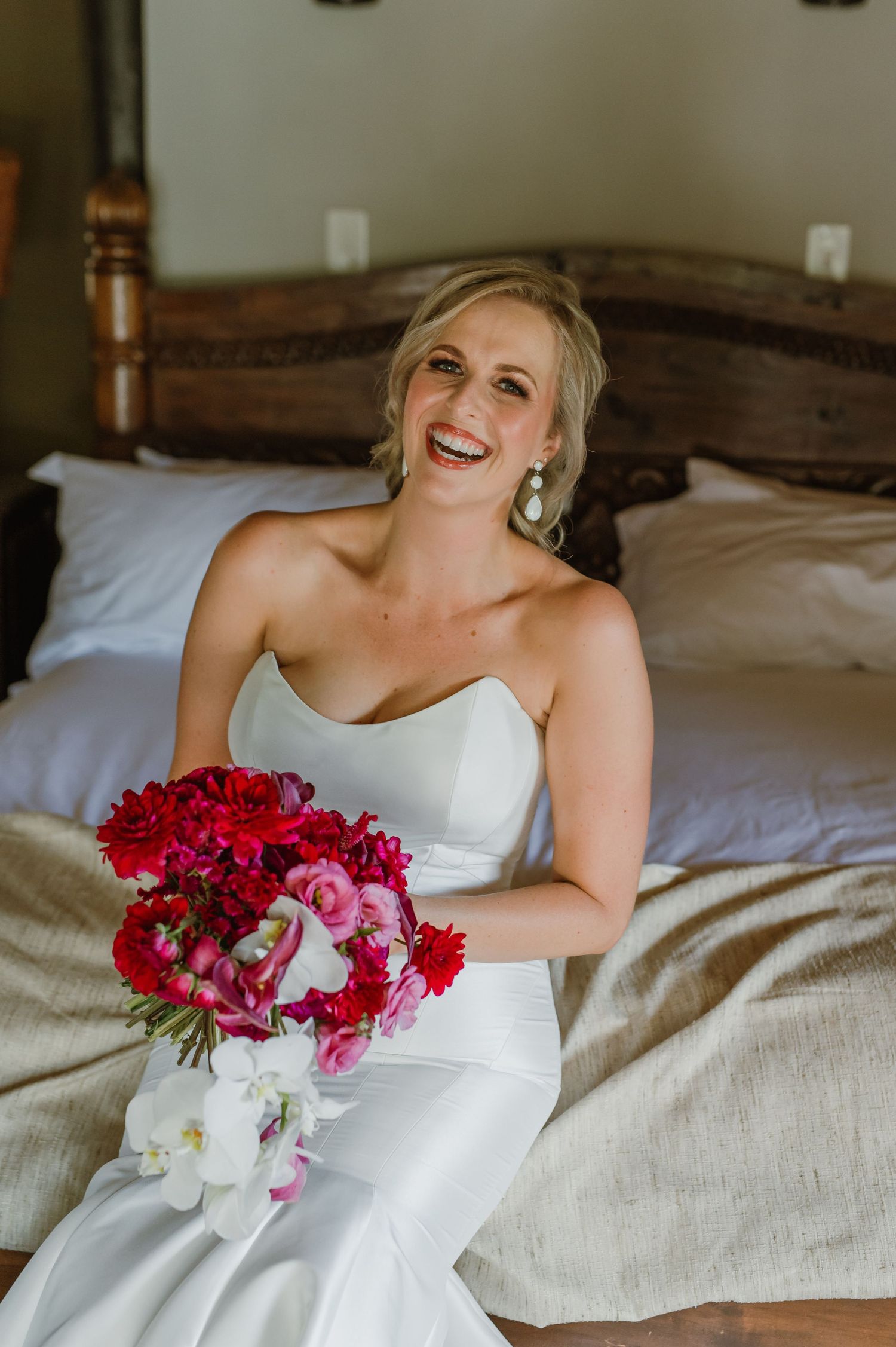 Elegant bridal portrait on a bed with vibrant red and pink floral bouquet.