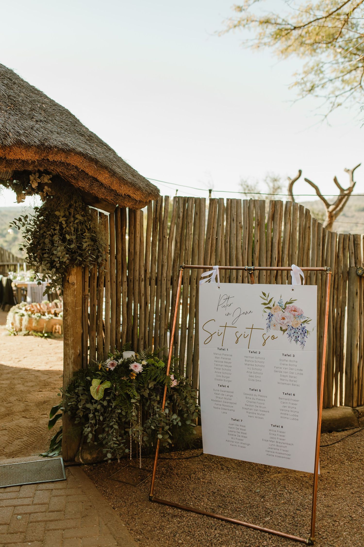 Wedding seating chart display with floral decorations against rustic wooden fence and thatched roof structure.