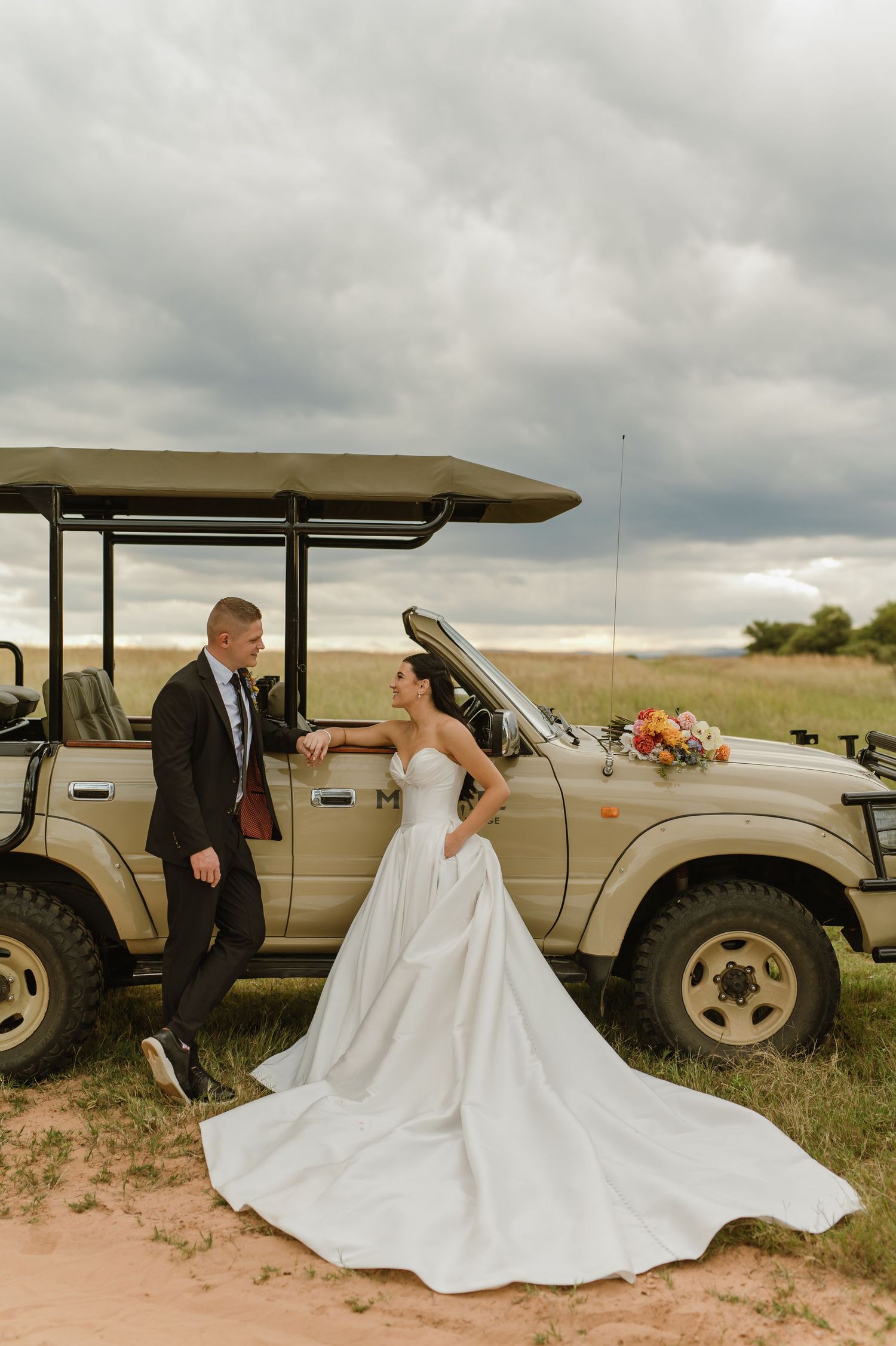 Bride in stunning white gown and groom in dark suit pose beside safari jeep in African savanna.