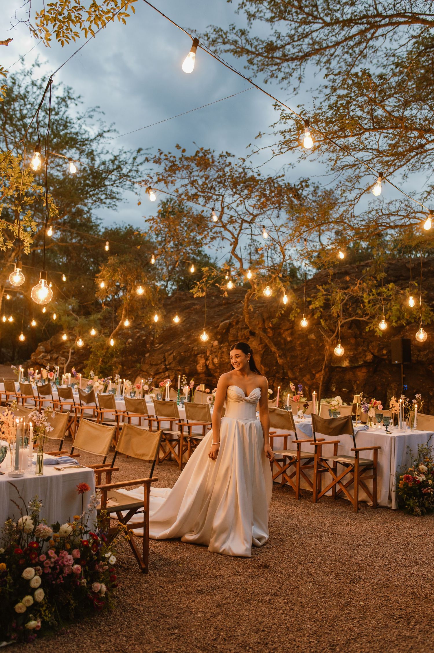 Bride in strapless white gown stands under warm string lights at beautifully set outdoor wedding venue.