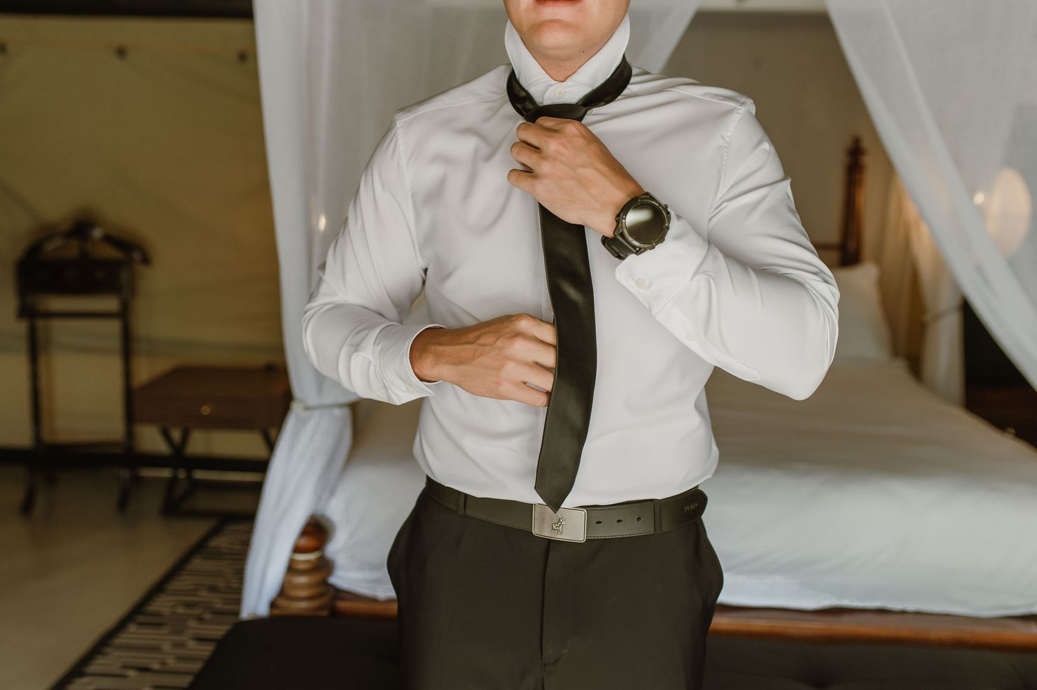 Groom in white dress shirt adjusting dark tie while getting ready in elegant bedroom.