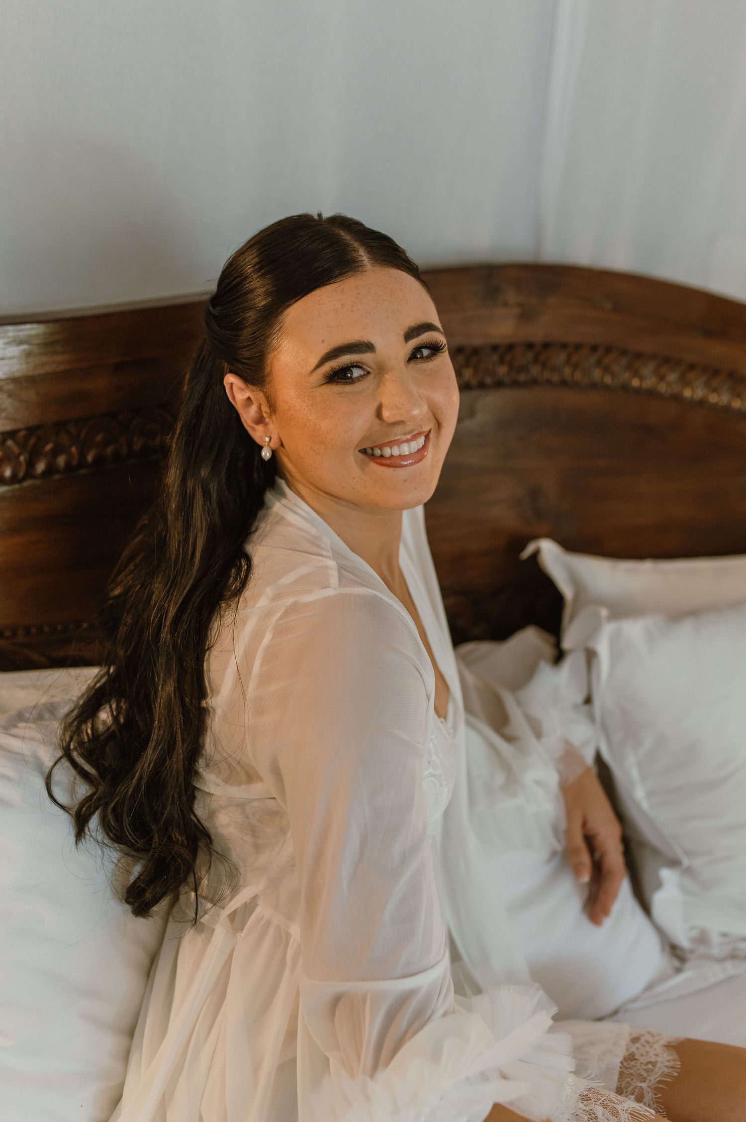 Smiling dark-haired bride in white sheer robe sitting on bed with carved wooden headboard.