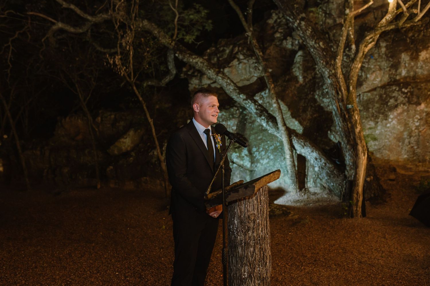 Man in black suit speaking at a rustic log podium during an outdoor nighttime event.