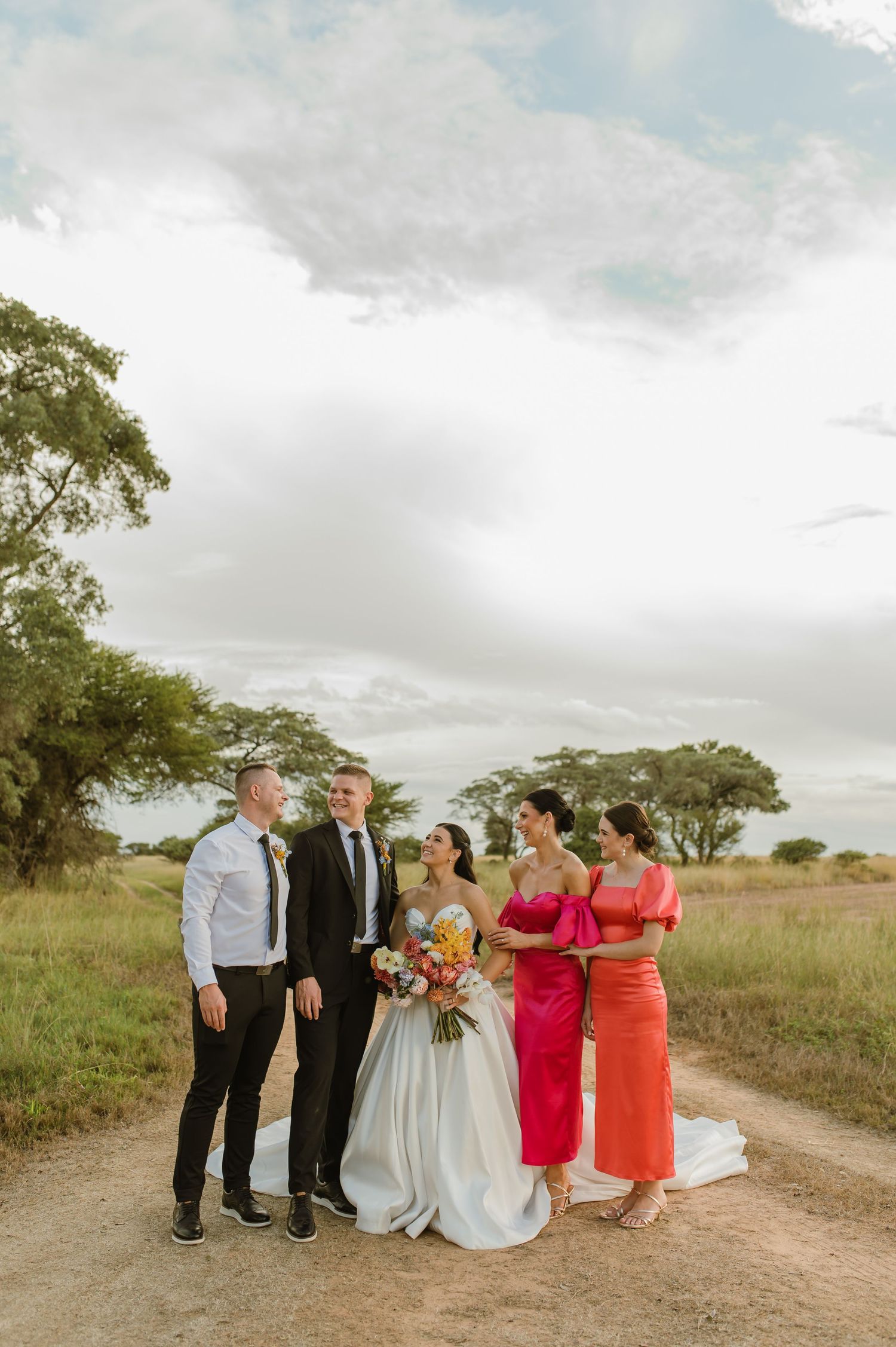 Wedding party posing on dirt road with bride, groom, and bridesmaids in pink and orange dresses.