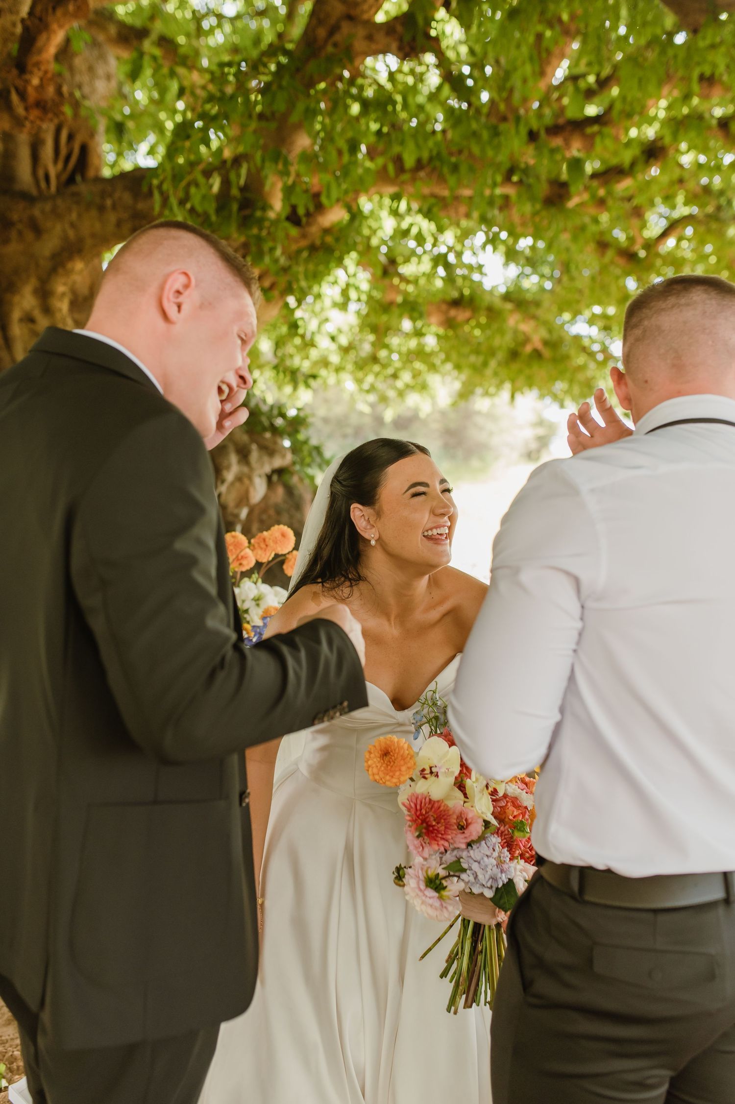 Bride laughing joyfully while holding bouquet during outdoor wedding ceremony under large tree.