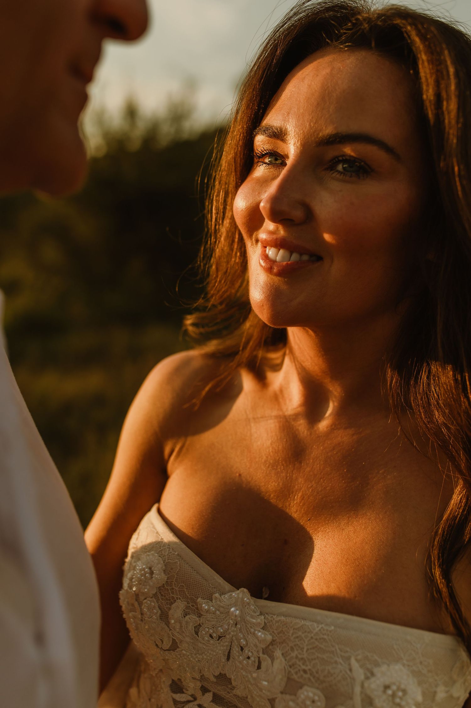 Bride in lace off-shoulder gown smiles warmly at golden hour, bathed in warm sunlight with green trees behind.