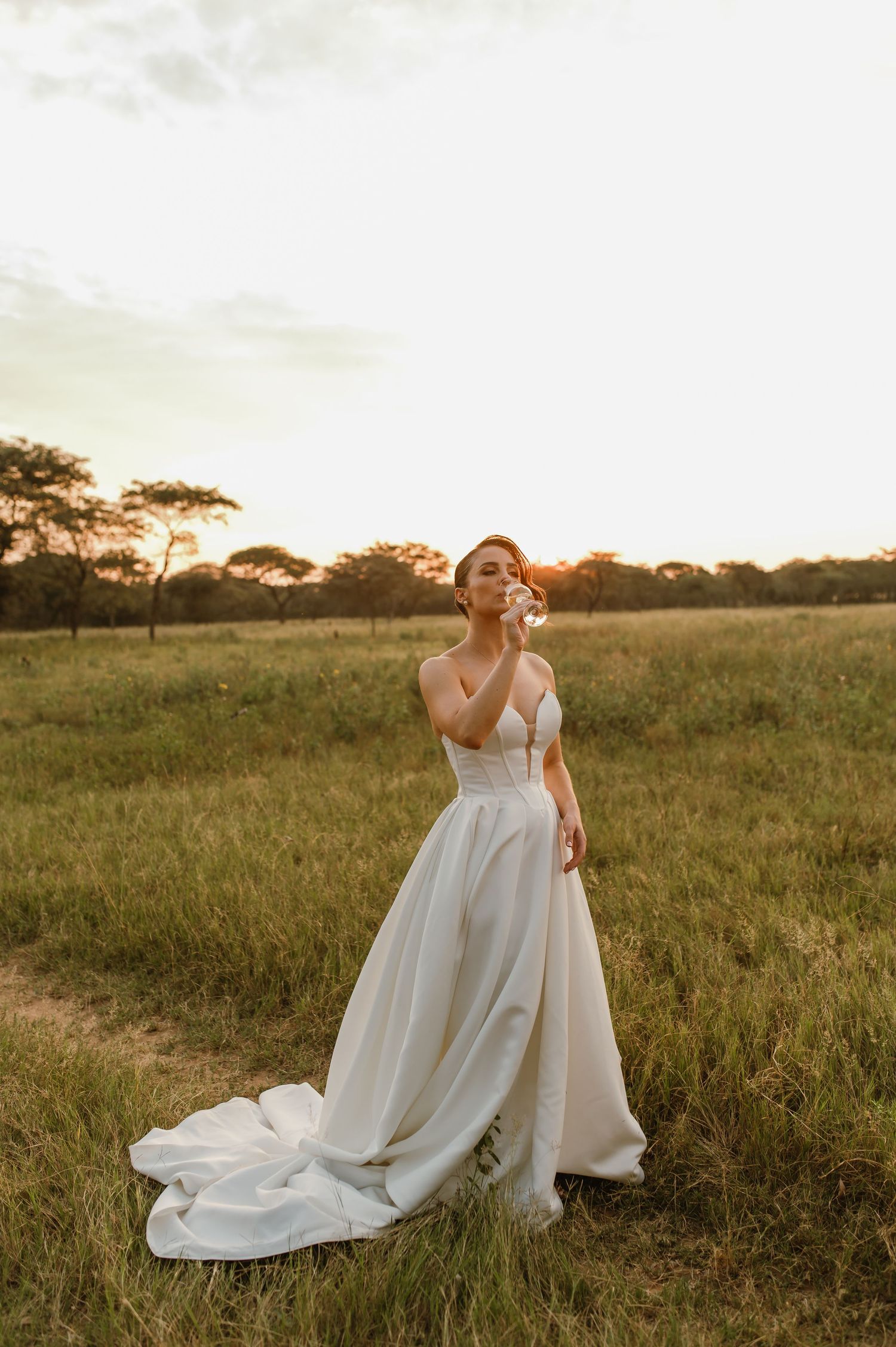 A bride in a flowing white wedding gown poses in a golden-lit field at sunset with acacia trees in the background.