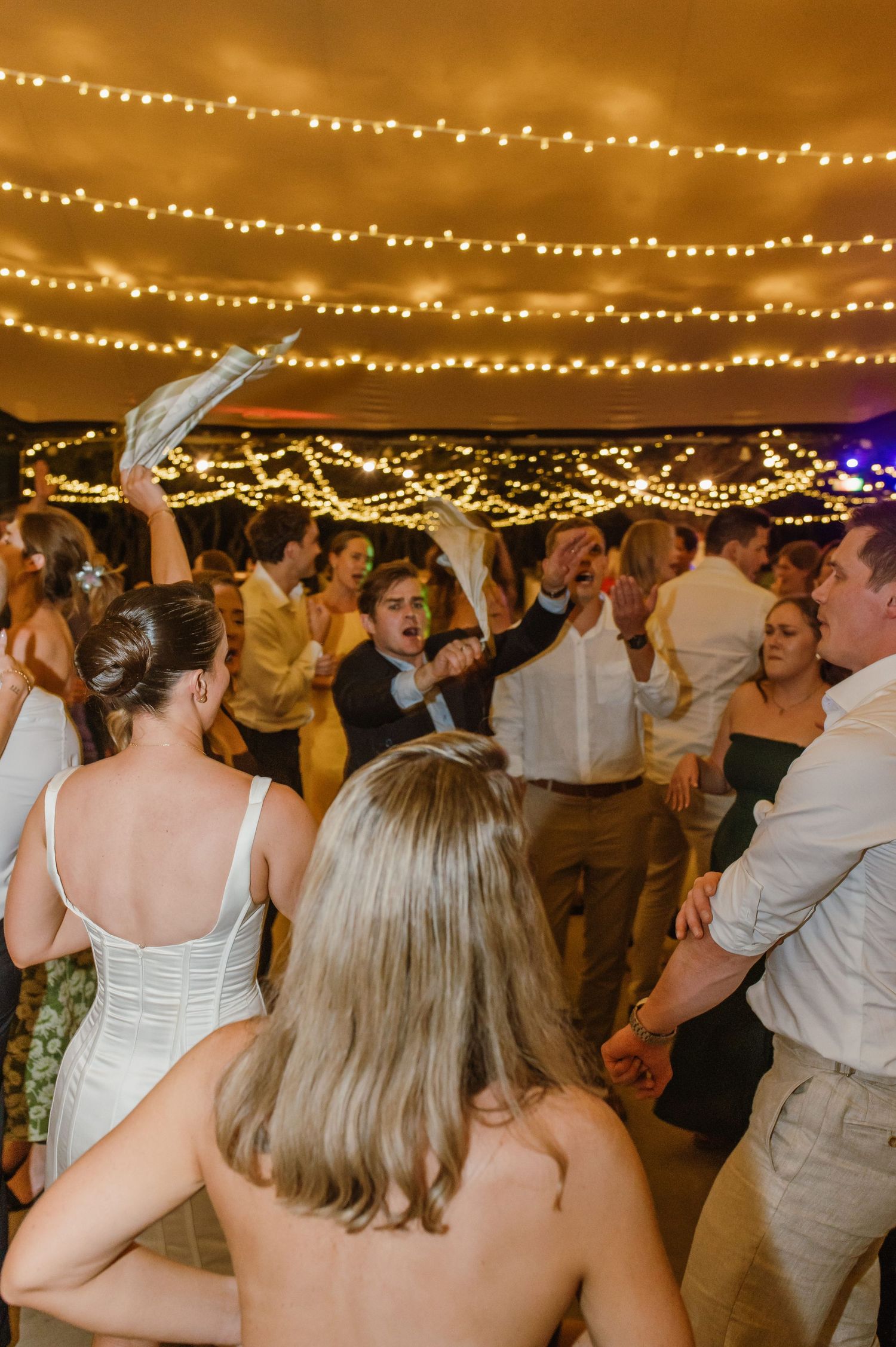 Wedding reception dance floor filled with guests celebrating under warm string light canopy.
