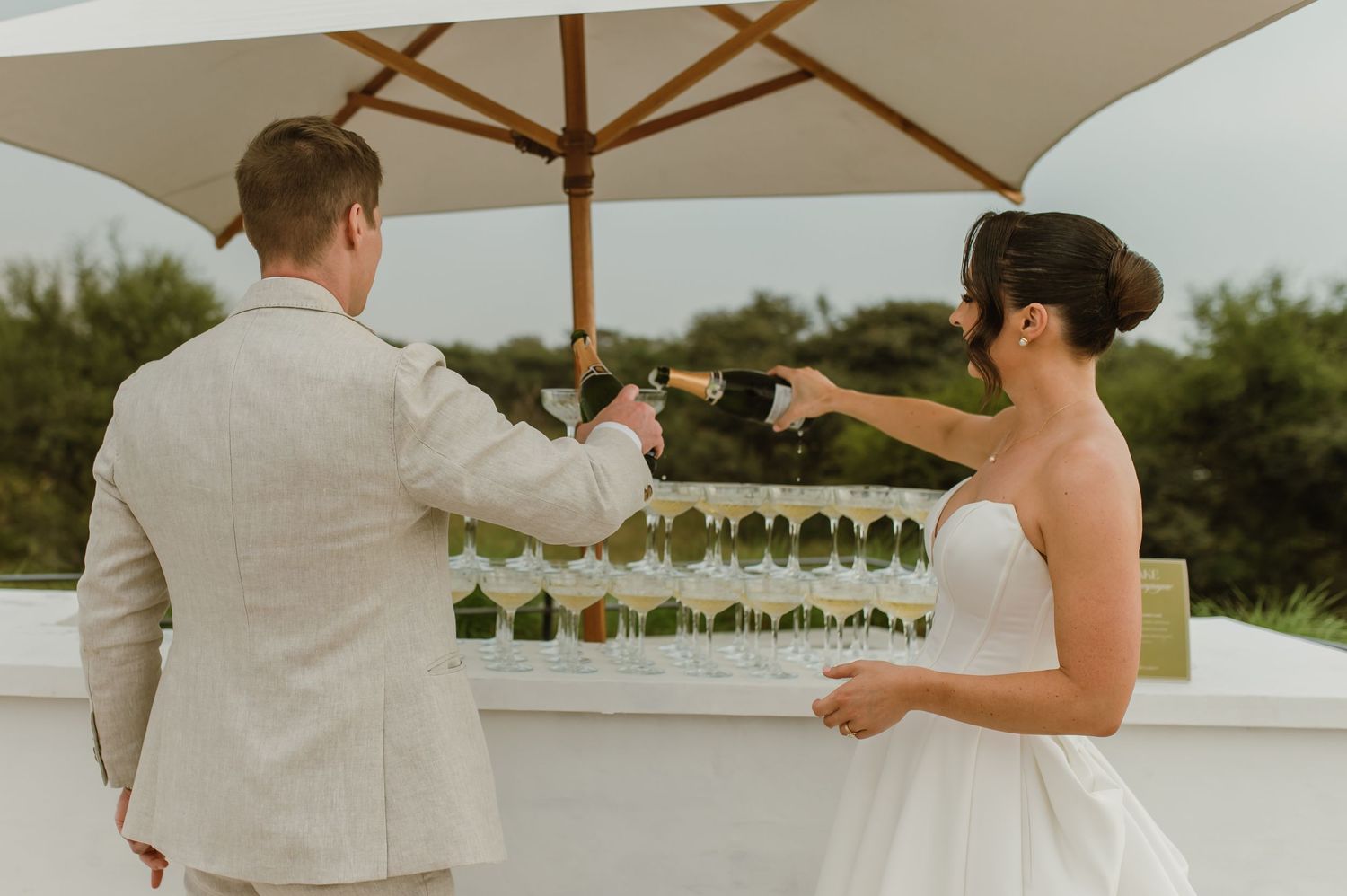 A couple in wedding attire toasts with champagne glasses under a white umbrella on an outdoor patio.