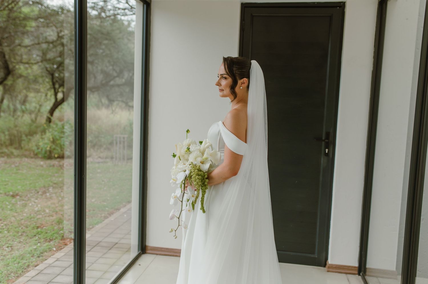 Side profile of bride in classic white wedding gown holding bouquet by window.