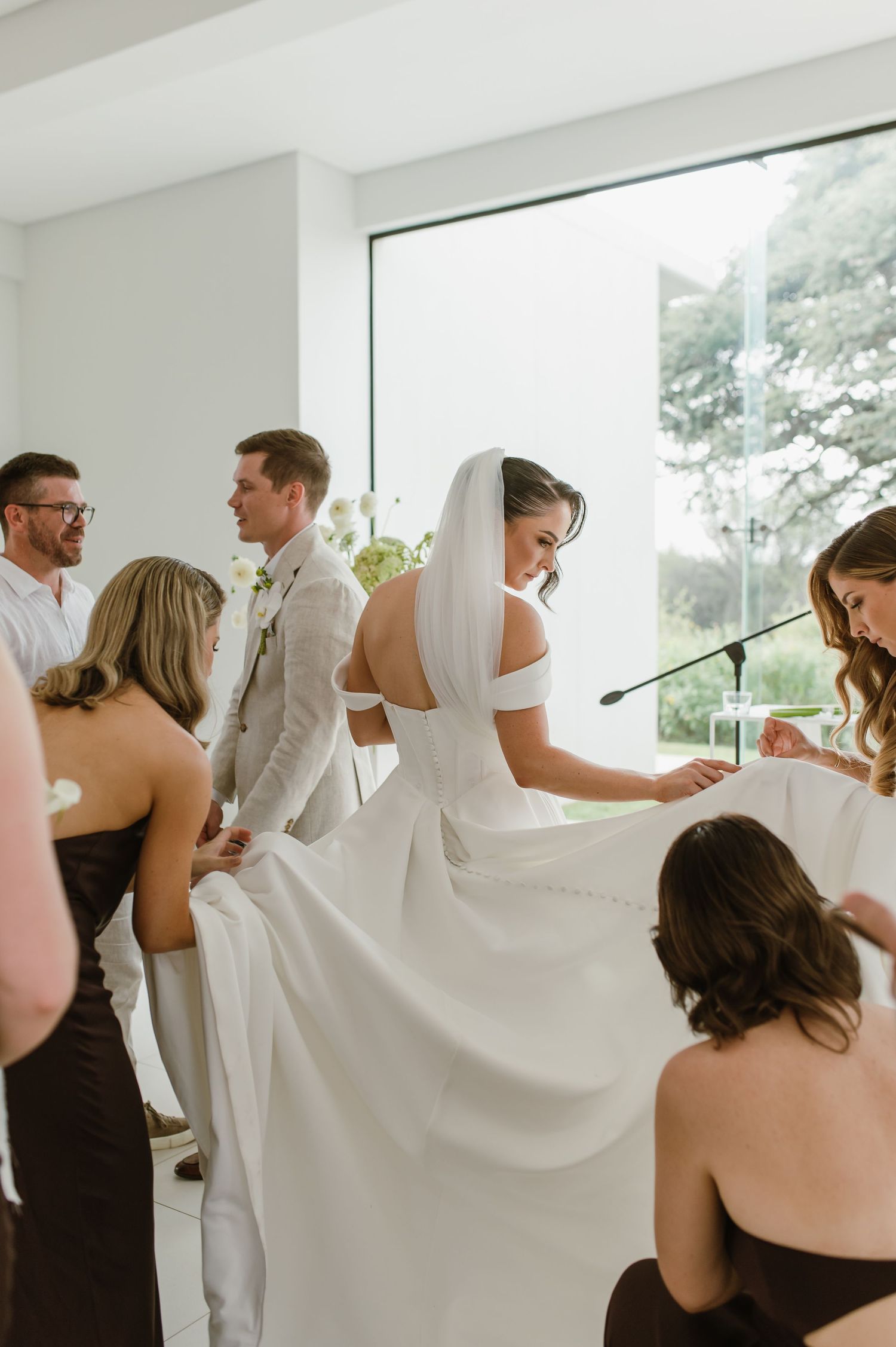 Wedding party members help arrange a flowing white wedding dress in a bright modern space.