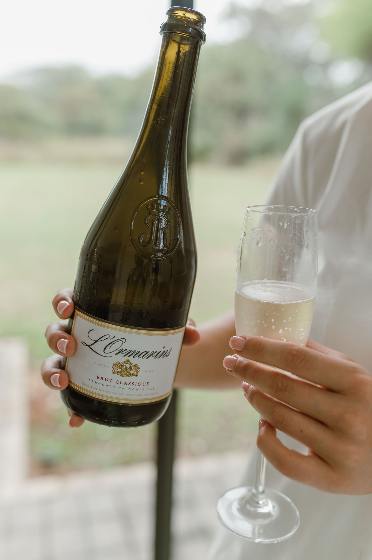 Hand holding bottle of L'Orangerie wine and champagne glass against blurred outdoor background.