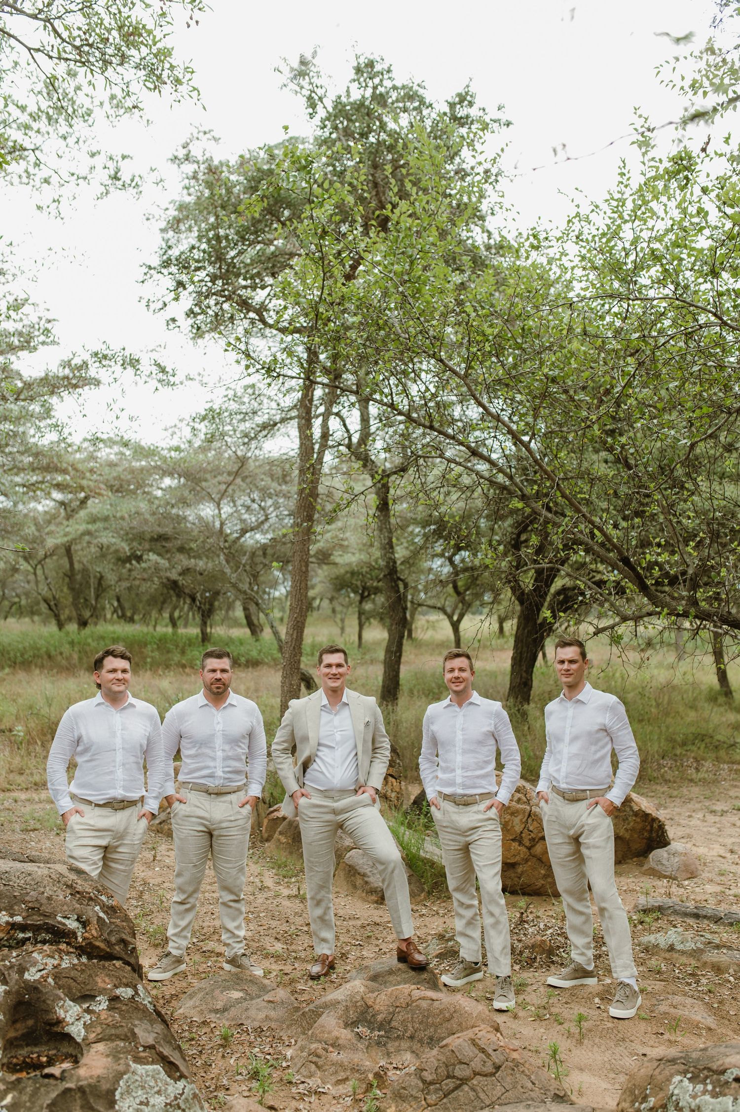 Five people in coordinated white and beige attire pose together on rocky terrain with trees in the background.