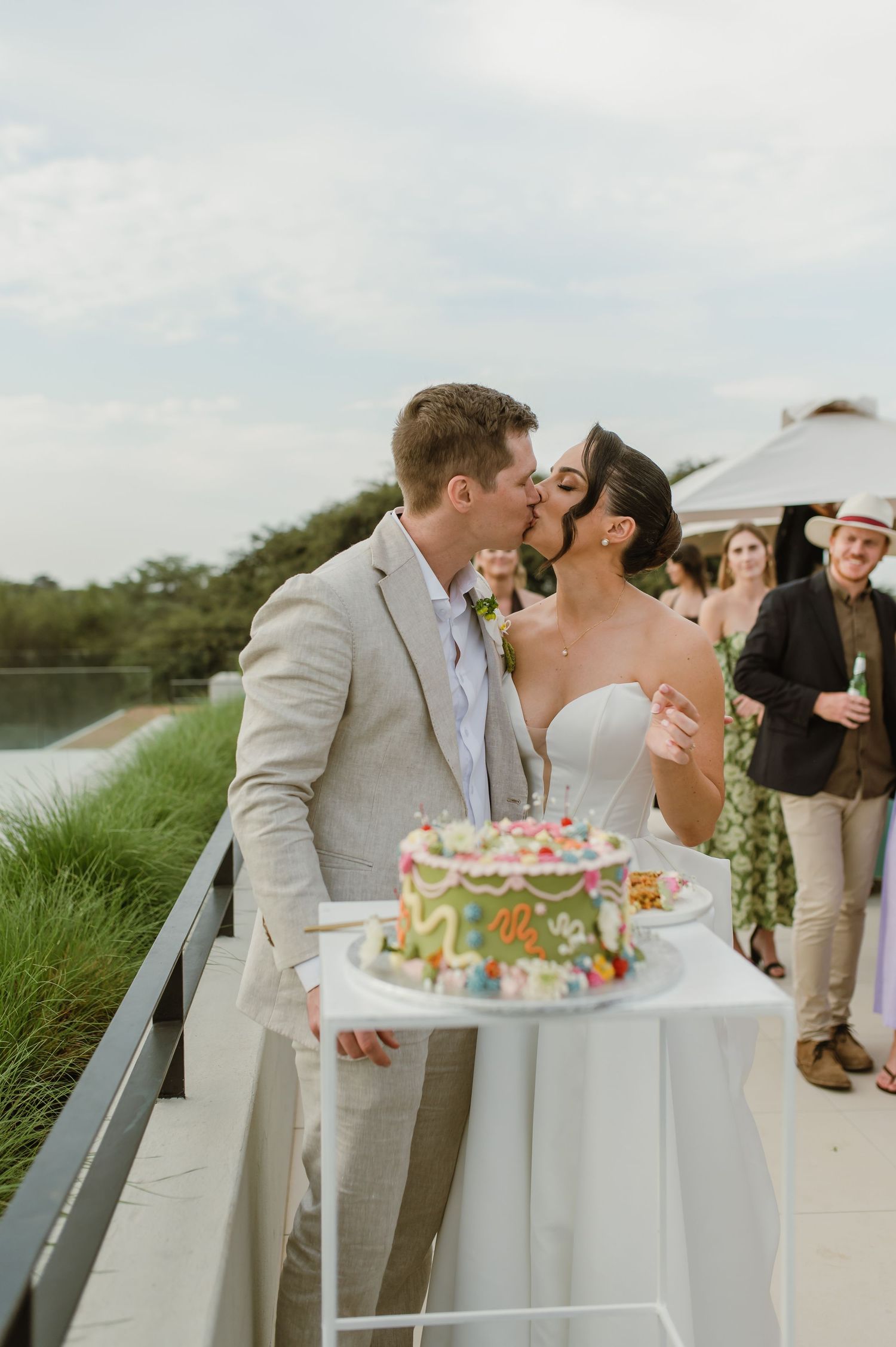 A couple shares a romantic kiss during their beach wedding cake cutting ceremony with guests watching.