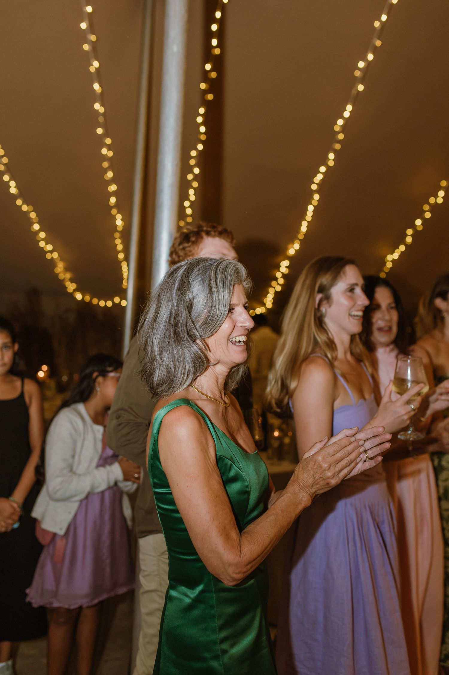 Wedding guests in formal attire clapping and celebrating under warm string lights.