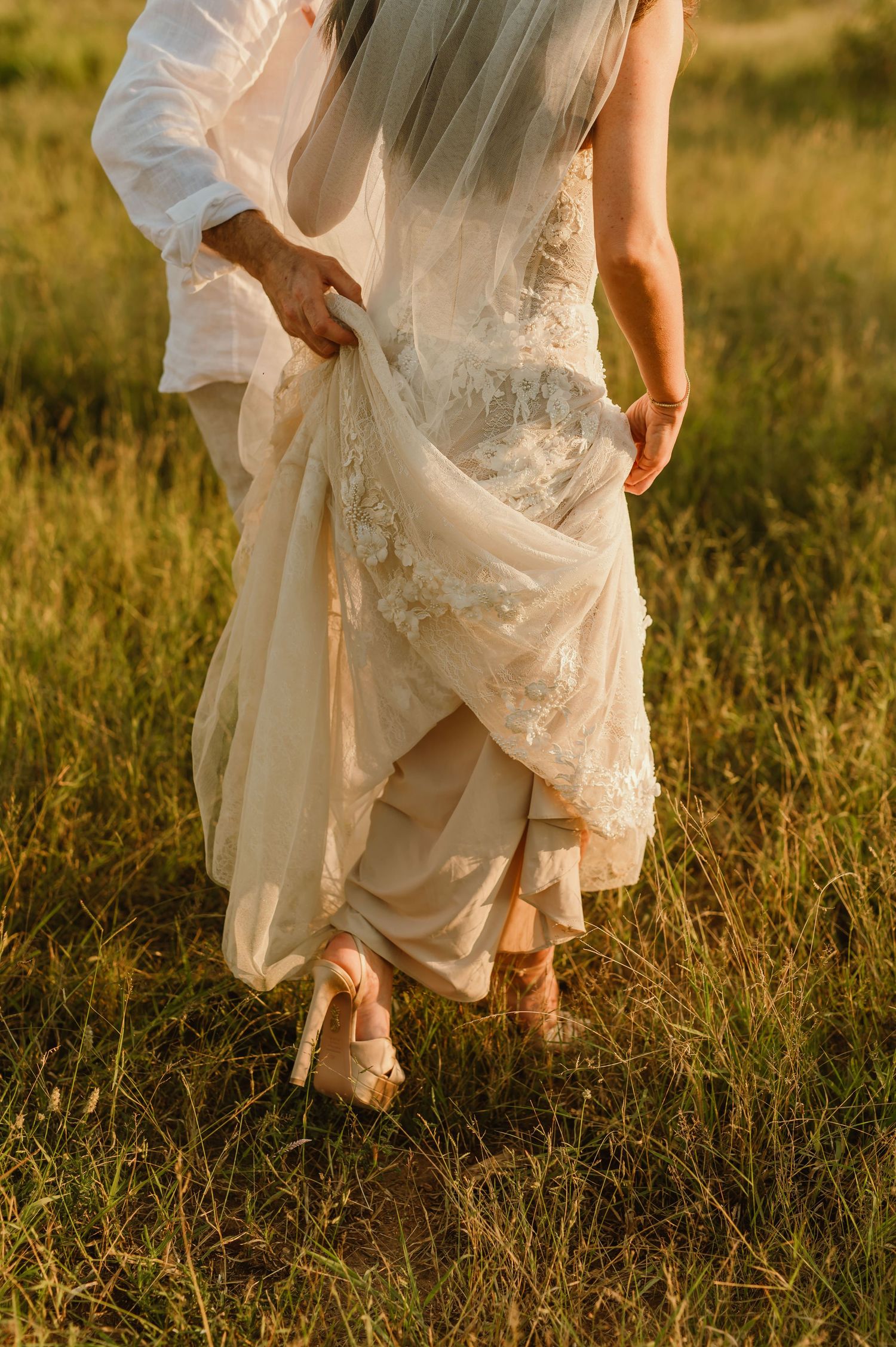 Bride in lace wedding gown and veil walks through golden grass field holding dress hem with groom in white shirt.