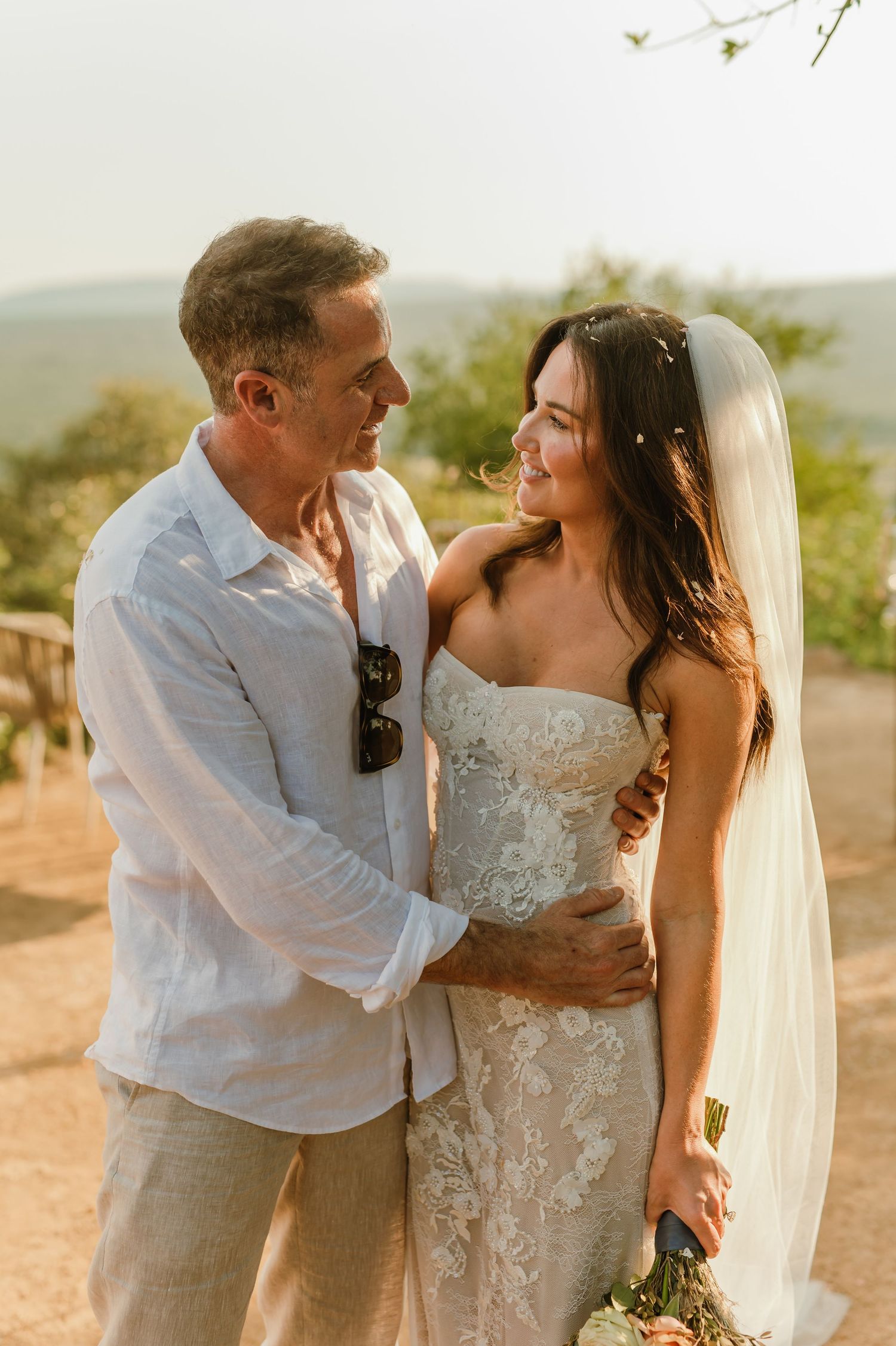 Bride in lace strapless gown and veil embraces groom in linen shirt outdoors at golden hour.