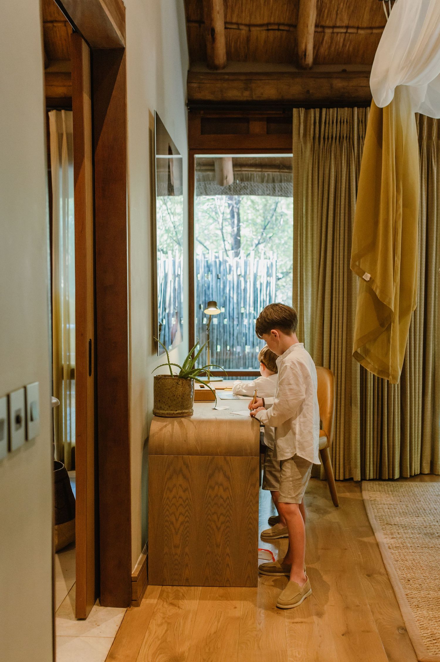 Boy in white shirt standing at wooden desk in luxury safari lodge room with large window and natural decor.