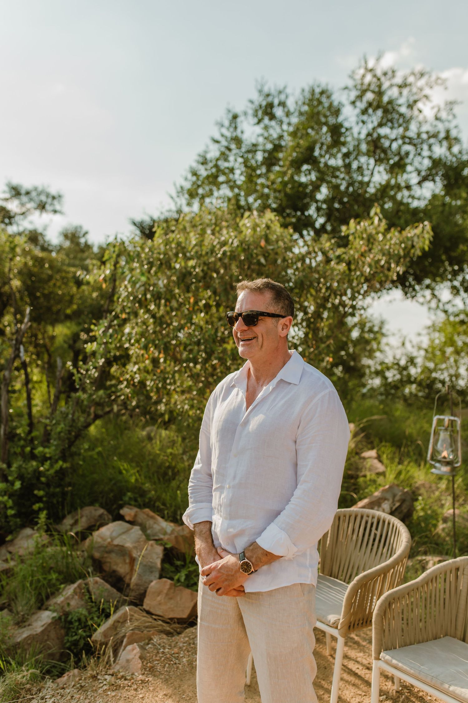 Smiling man in white linen shirt and sunglasses standing outdoors at a garden wedding venue.