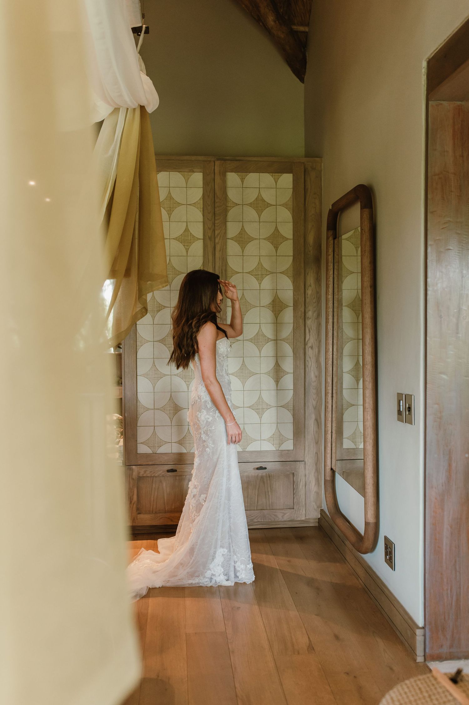 Bride in elegant white lace wedding gown standing in a rustic wooden room with decorative cabinet and floor mirror.