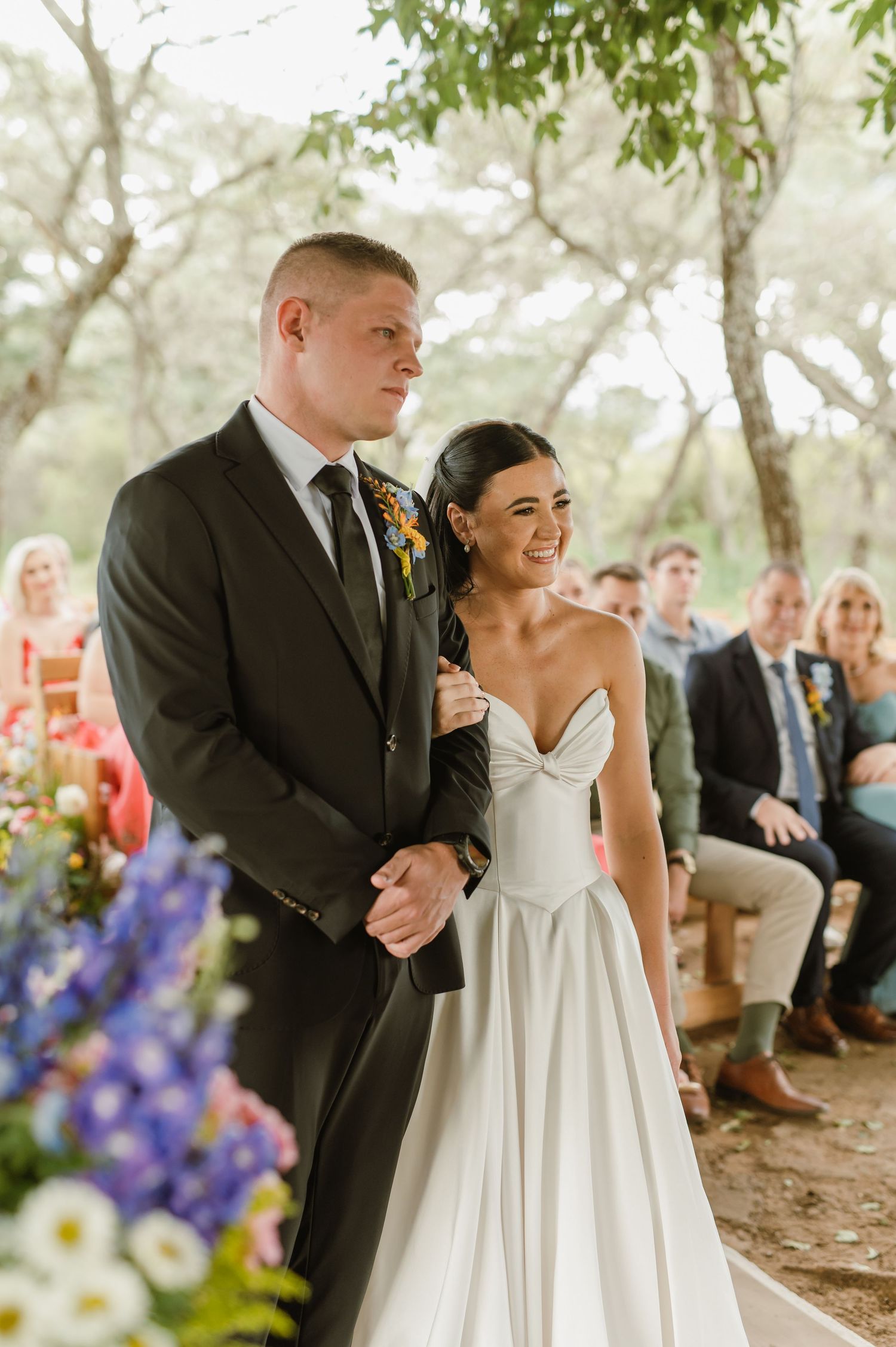 Bride smiles at groom in dark suit during outdoor wedding ceremony among trees.