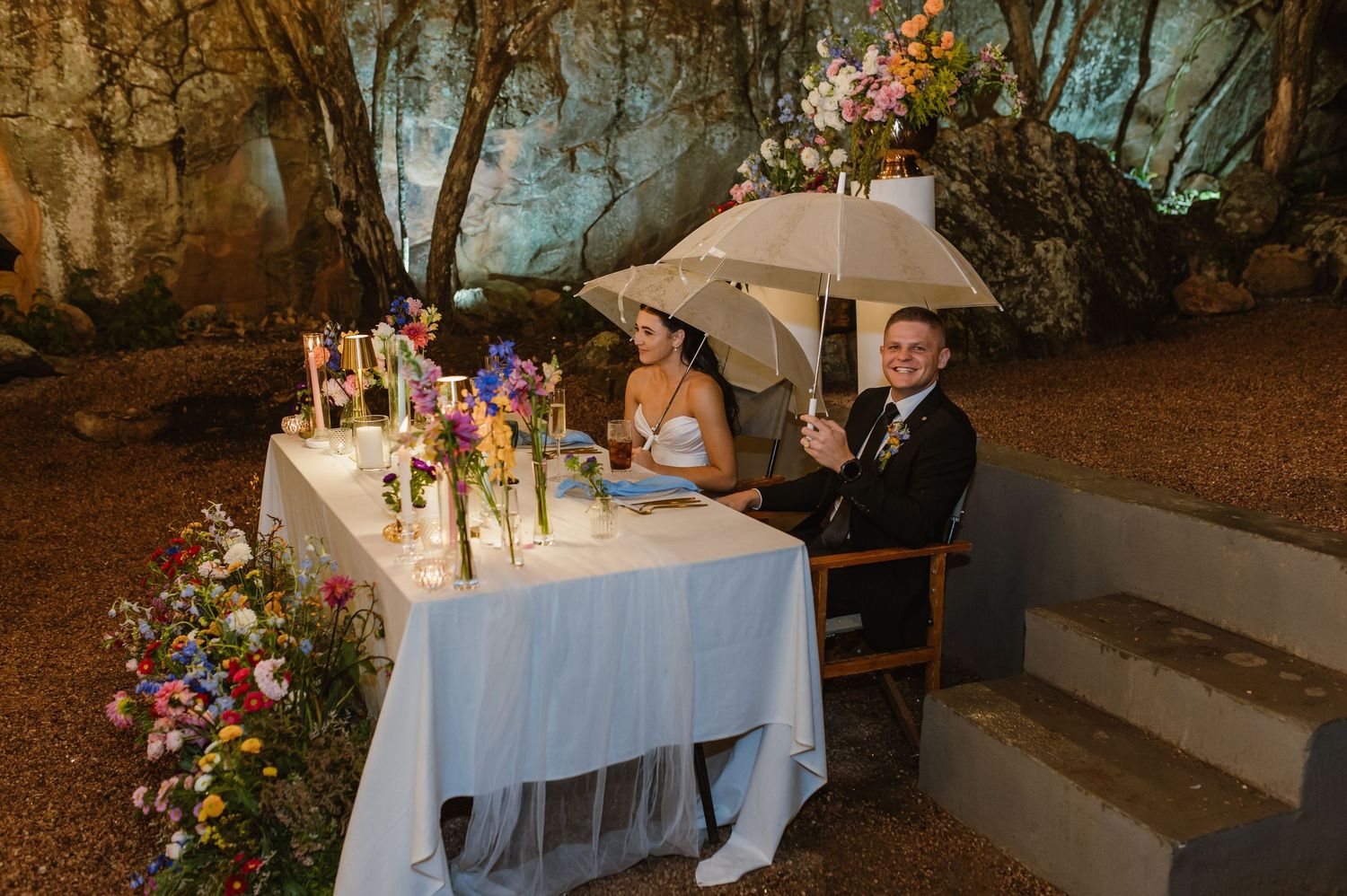 Bride and groom sit at sweetheart table under white umbrella in rustic cave wedding venue.