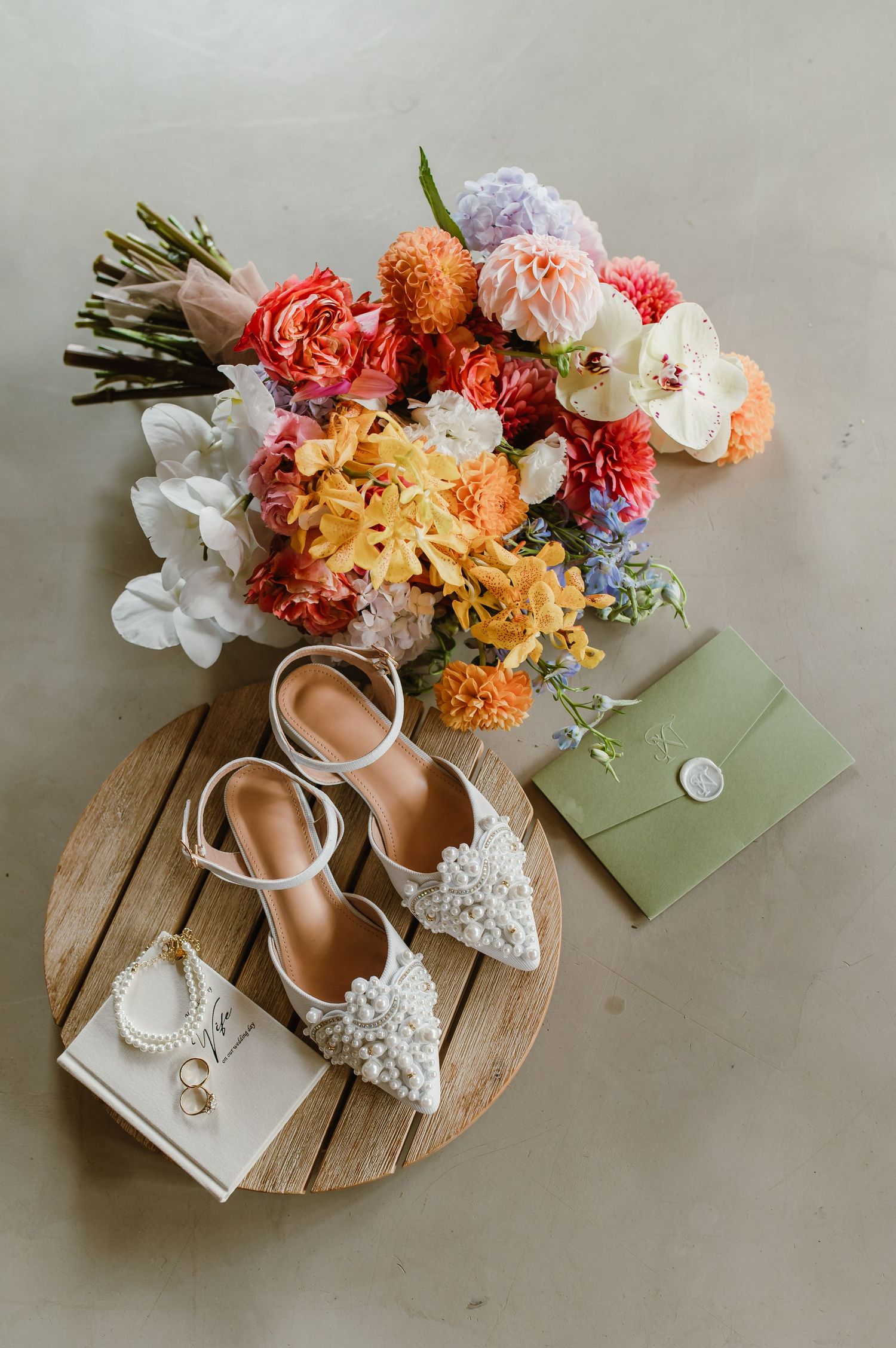 Colorful wedding bouquet with bridal heels, pearl jewelry, rings, and green envelope on wooden tray.