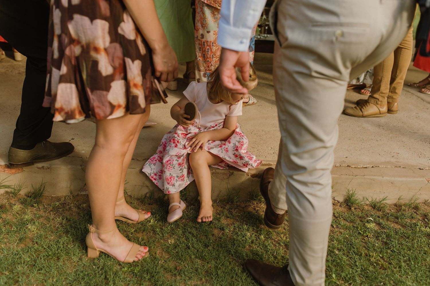 Young child in floral dress sitting on ground surrounded by wedding guests legs at outdoor ceremony.