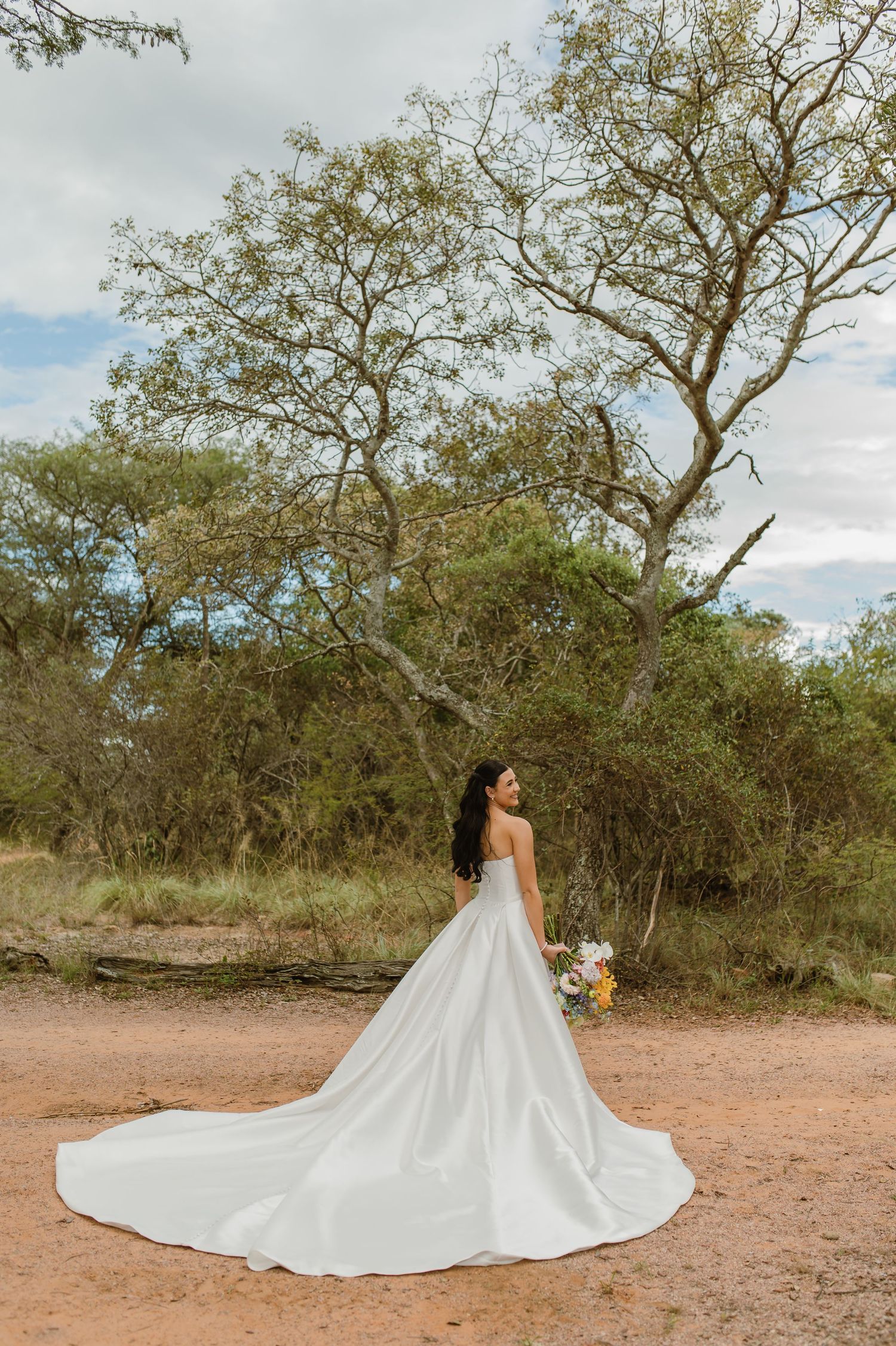 Bride in a stunning white ball gown with long train standing in a natural bush setting holding a bouquet.