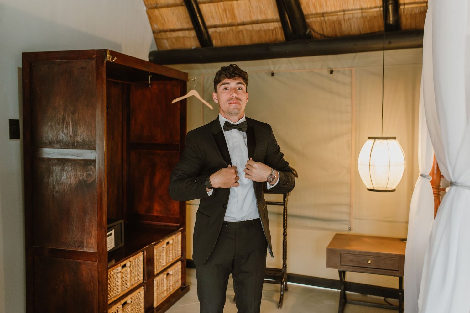 A formal portrait showing someone in a black bow tie and tuxedo getting ready in a rustic room.