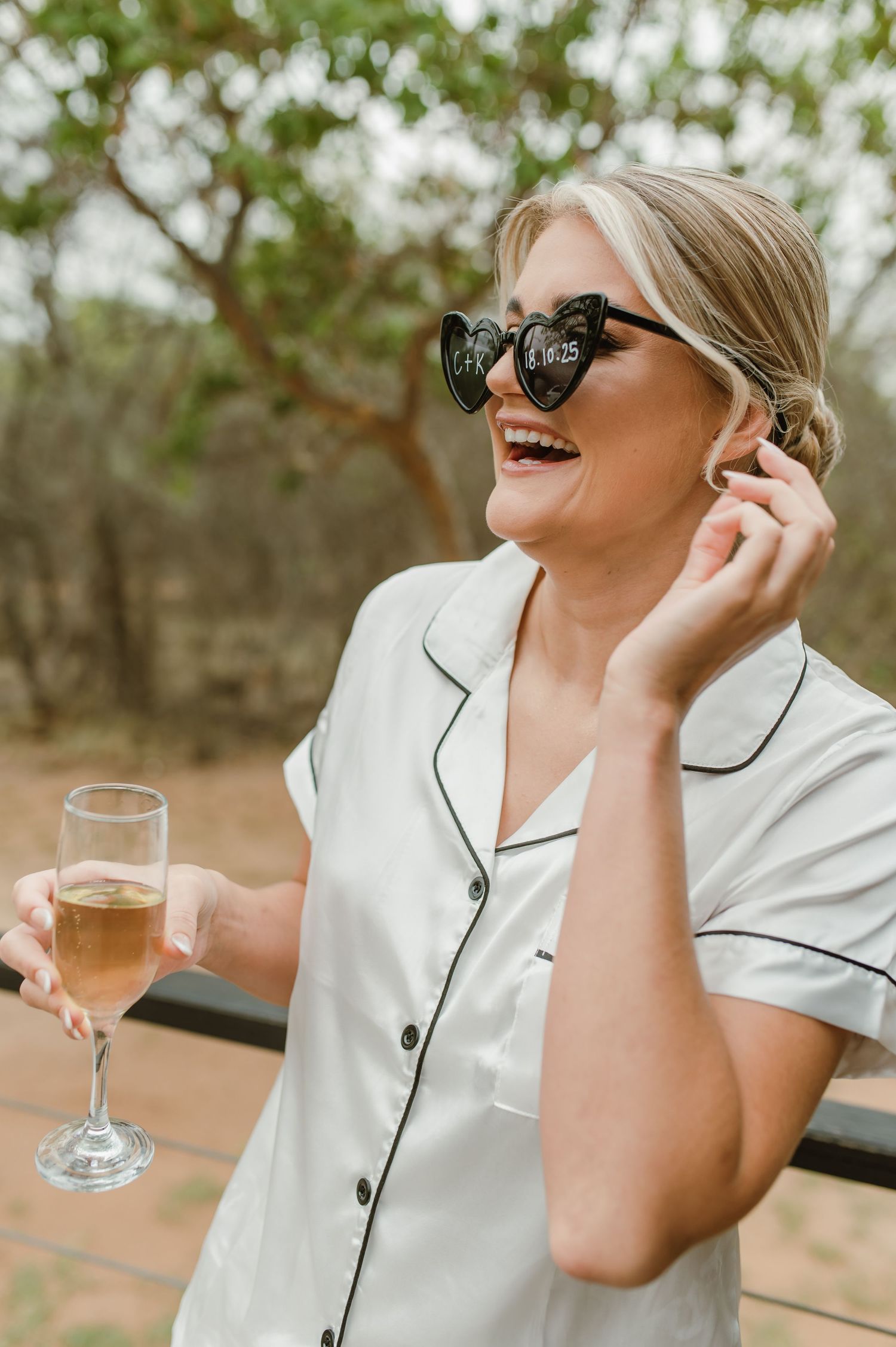 Person in white silk pajamas wearing sunglasses and holding champagne glass outdoors.