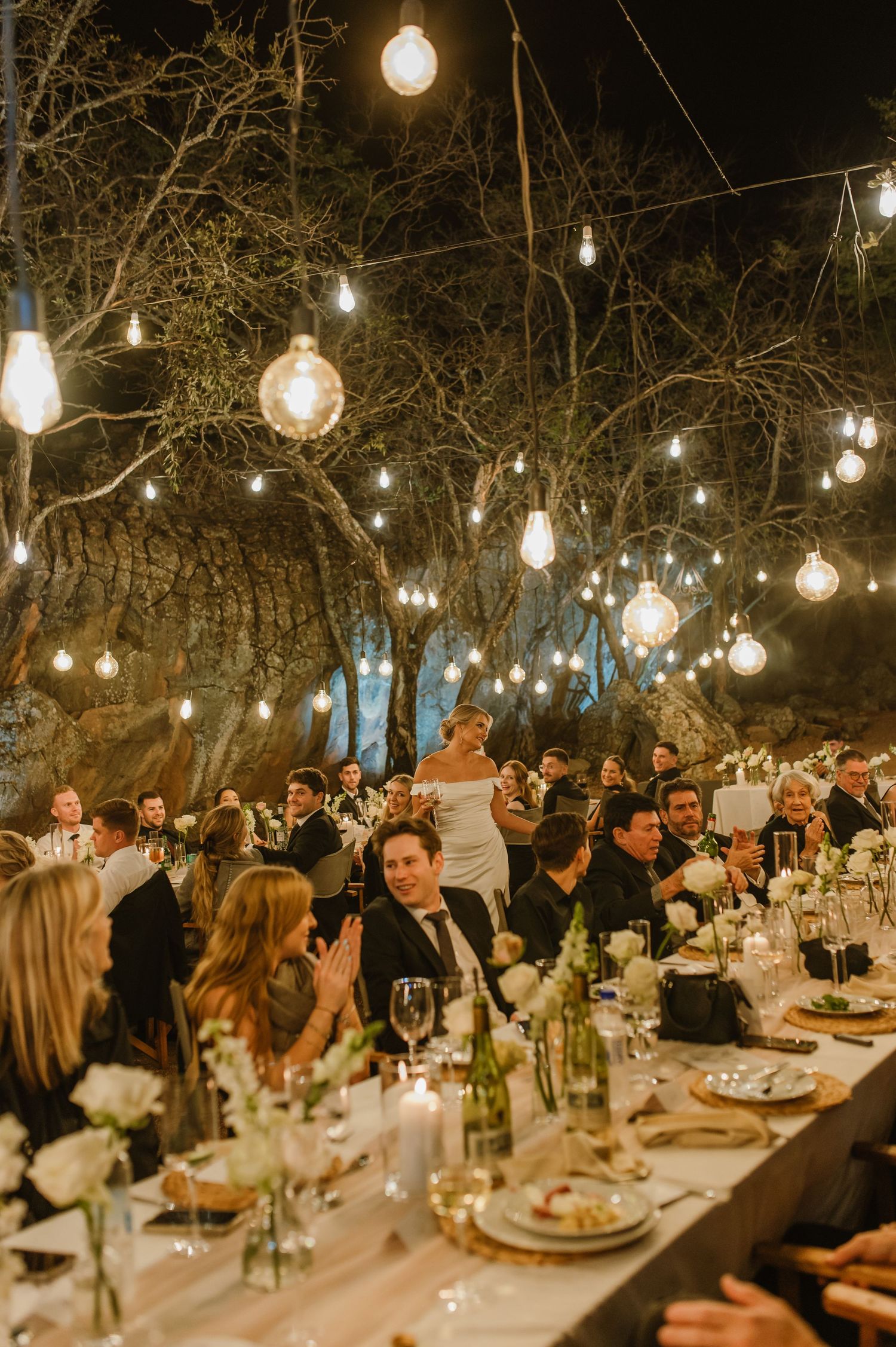 Candlelit wedding reception tables filled with guests under a canopy of string lights.